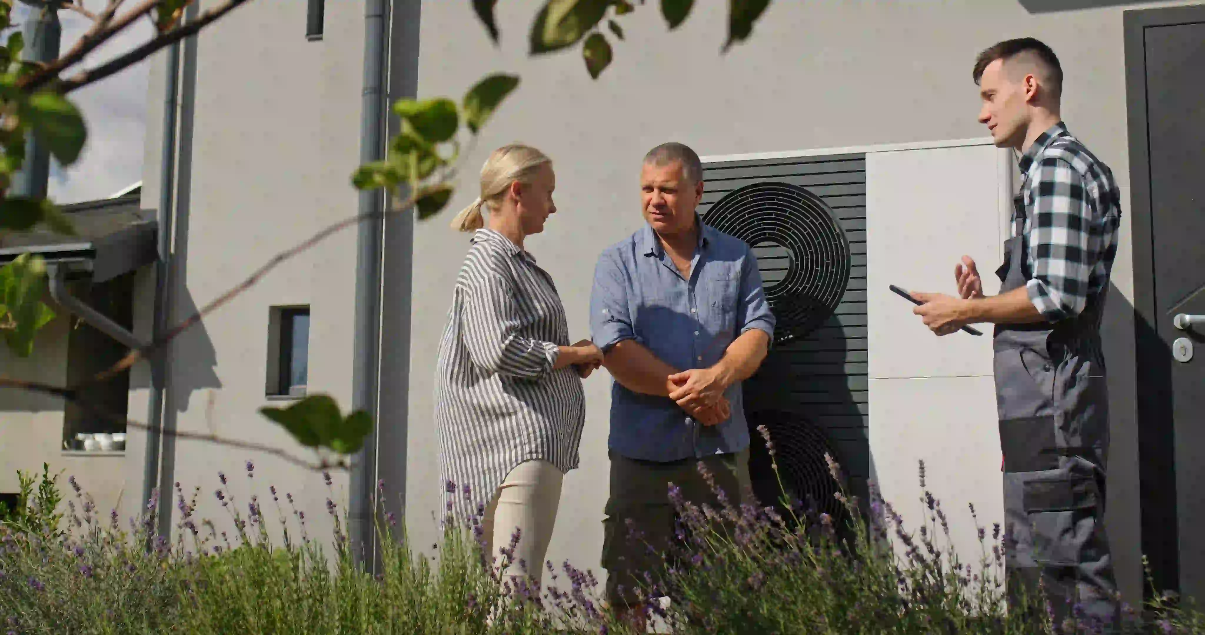 A technician in grey overalls, holding a tablet, speaks with a middle-aged couple standing in the yard of a modern home. A large heat pump is visible on the wall behind them. A blurred lavender bush and a leafy branch are in the foreground.