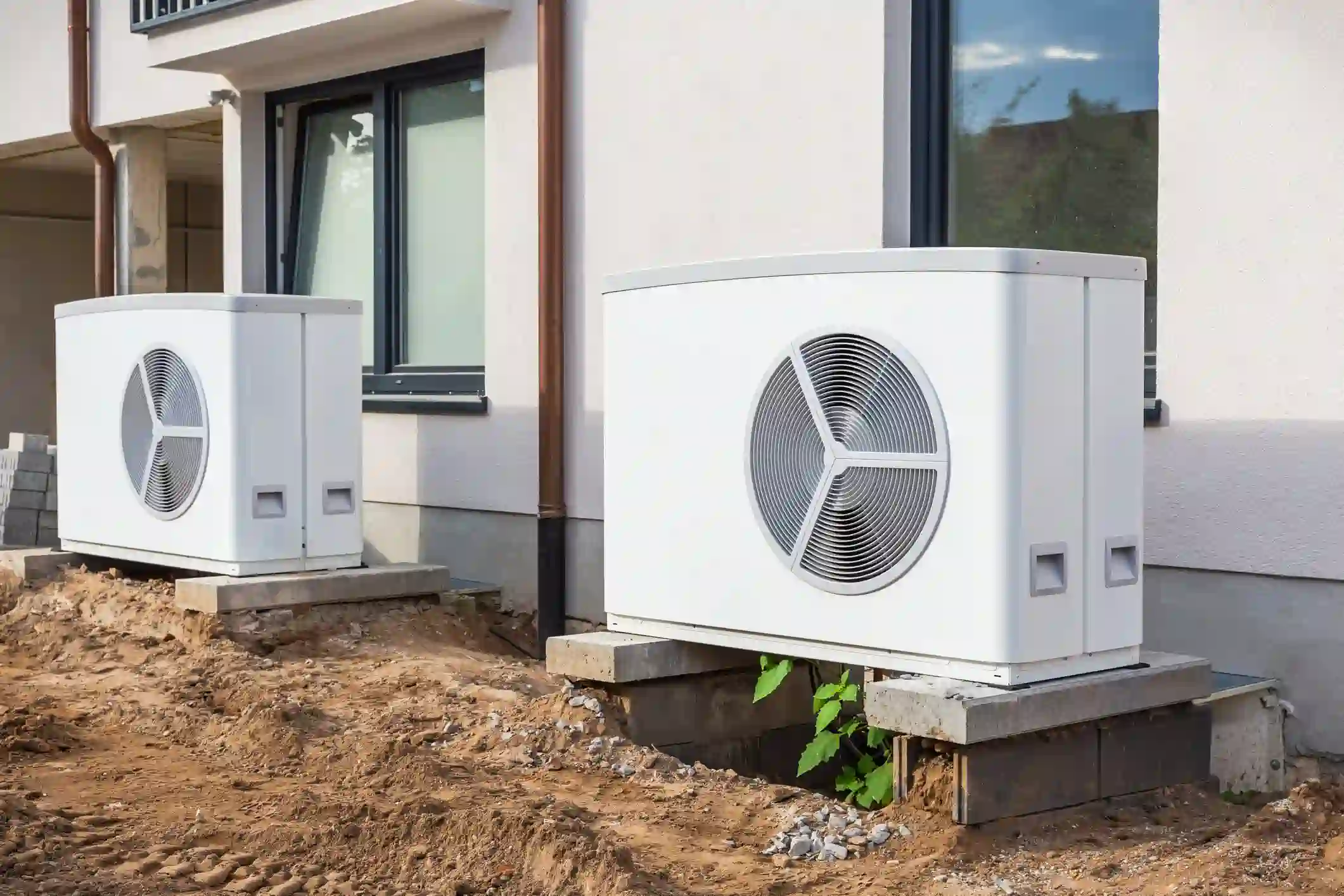 Two modern, rectangular heat pump units are installed side-by-side on concrete blocks outside a new house under construction. The ground is bare and muddy, with some small stones and weeds visible. The house has clean, light-colored siding and large windows, suggesting a new build or recent renovation.