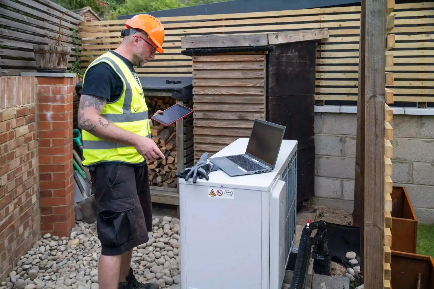 A technician, wearing an orange hard hat and a high-visibility vest, stands in a backyard. He is holding a tablet in one hand and looking down at an open laptop resting on top of a white heat pump unit. The yard features a brick wall, wooden fencing, and a patch of grey gravel.