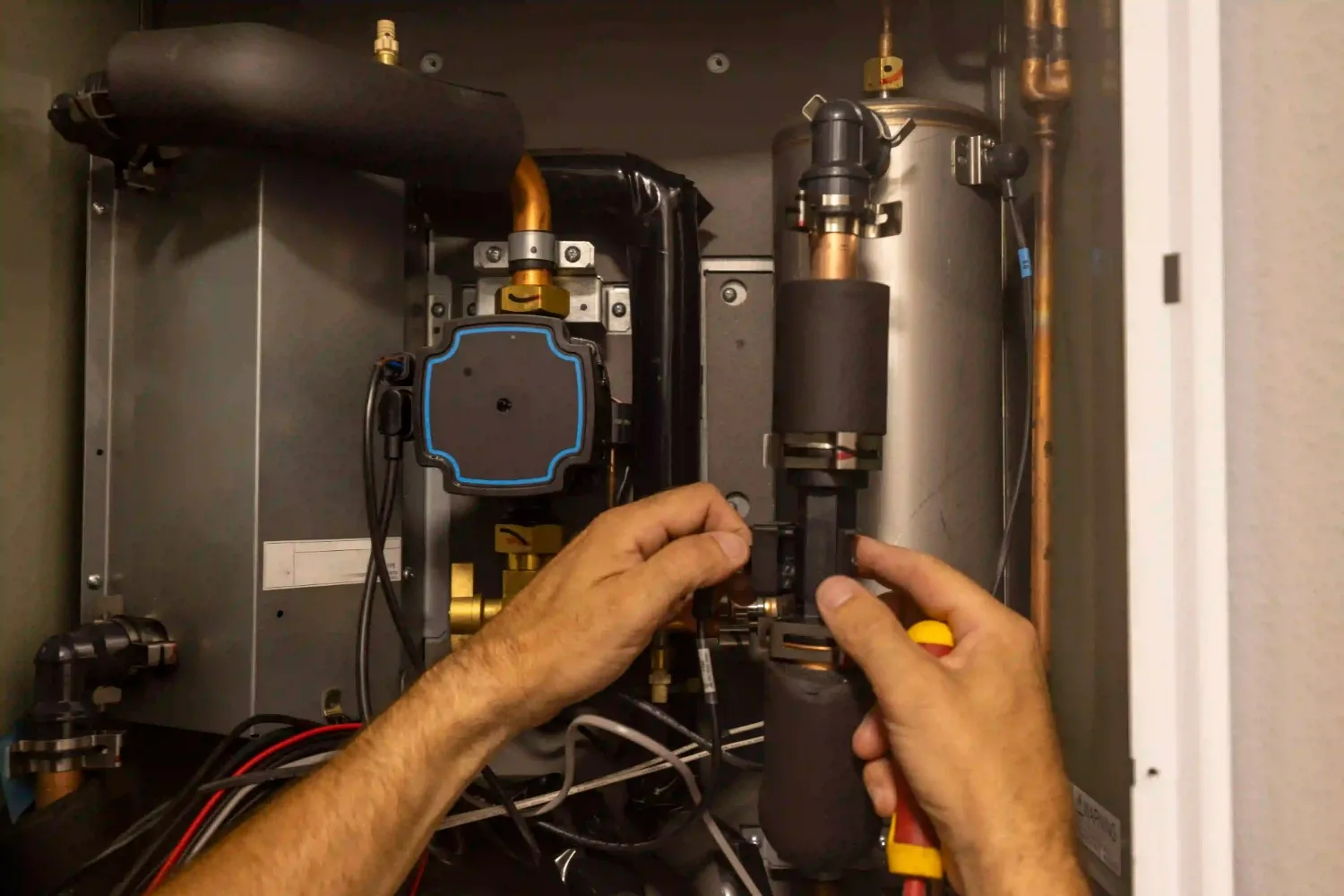 A pair of hands wearing black gloves is shown working on the internal components of a heat pump. The hands are holding and adjusting a part with a yellow and red tool. The surrounding area shows various pipes, wires, and a small pump.