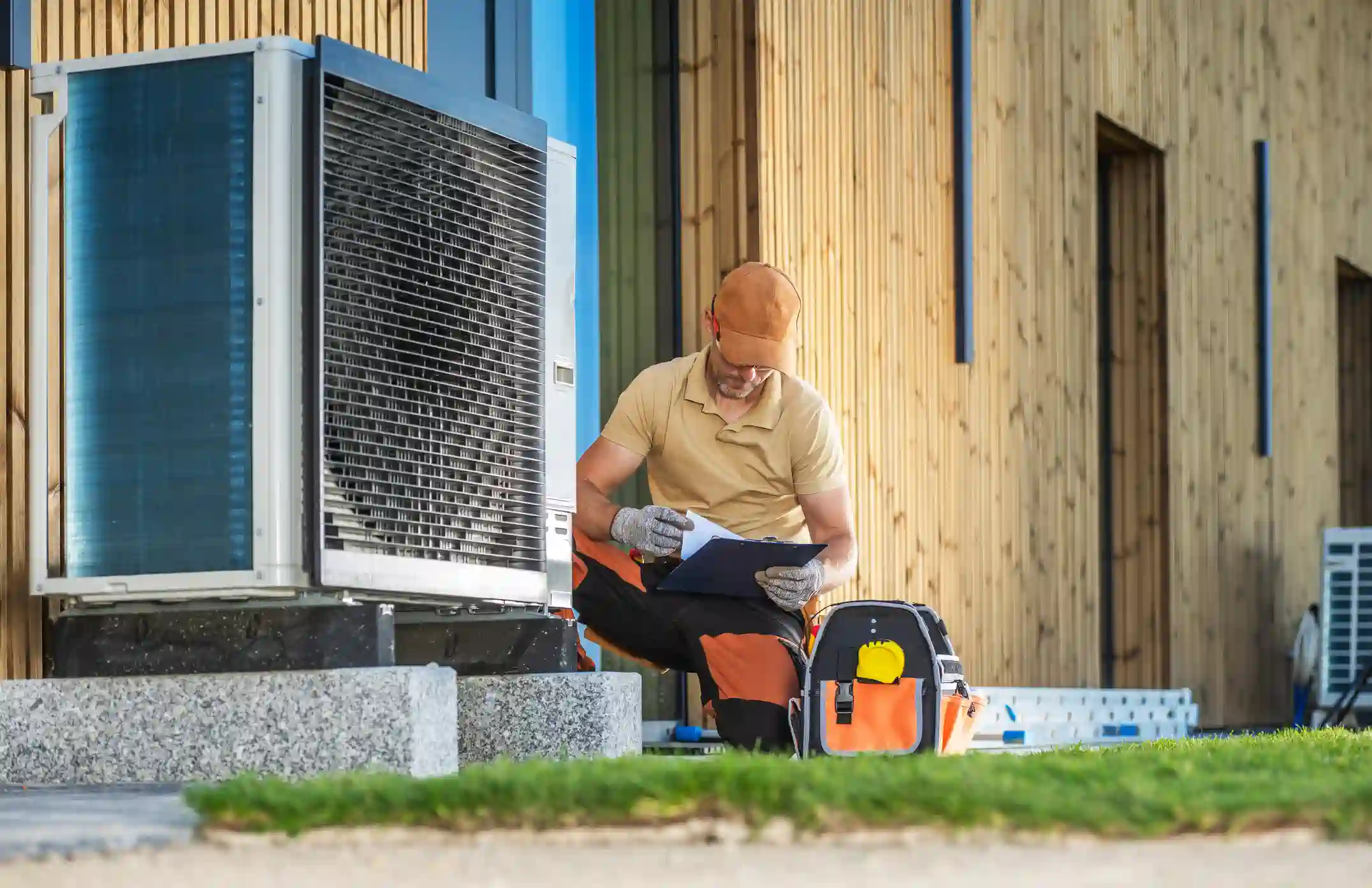 A male technician wearing a hat and gloves kneels by a large heat pump unit next to a modern house with wood siding. He is looking at a clipboard, with a bright orange and black tool bag beside him on the ground. A small section of green lawn is visible in the foreground.