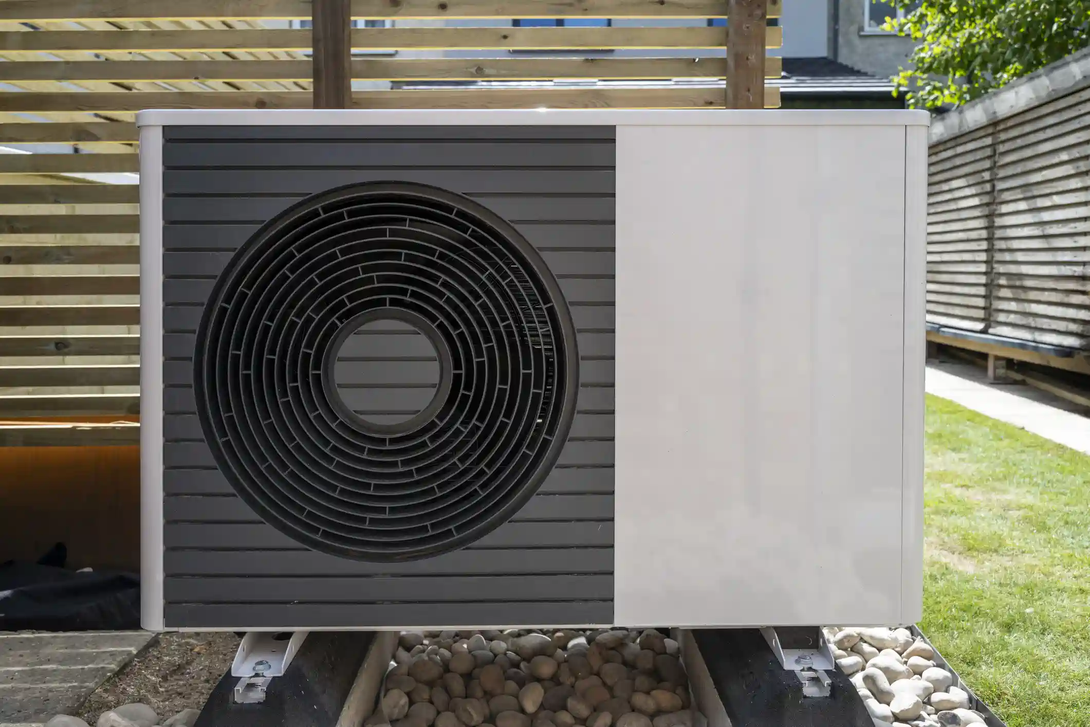 A close-up of a modern, white and gray heat pump unit with a circular fan grate on the left. The unit is installed on a bed of small, smooth rocks next to a wooden pergola and a grassy yard.