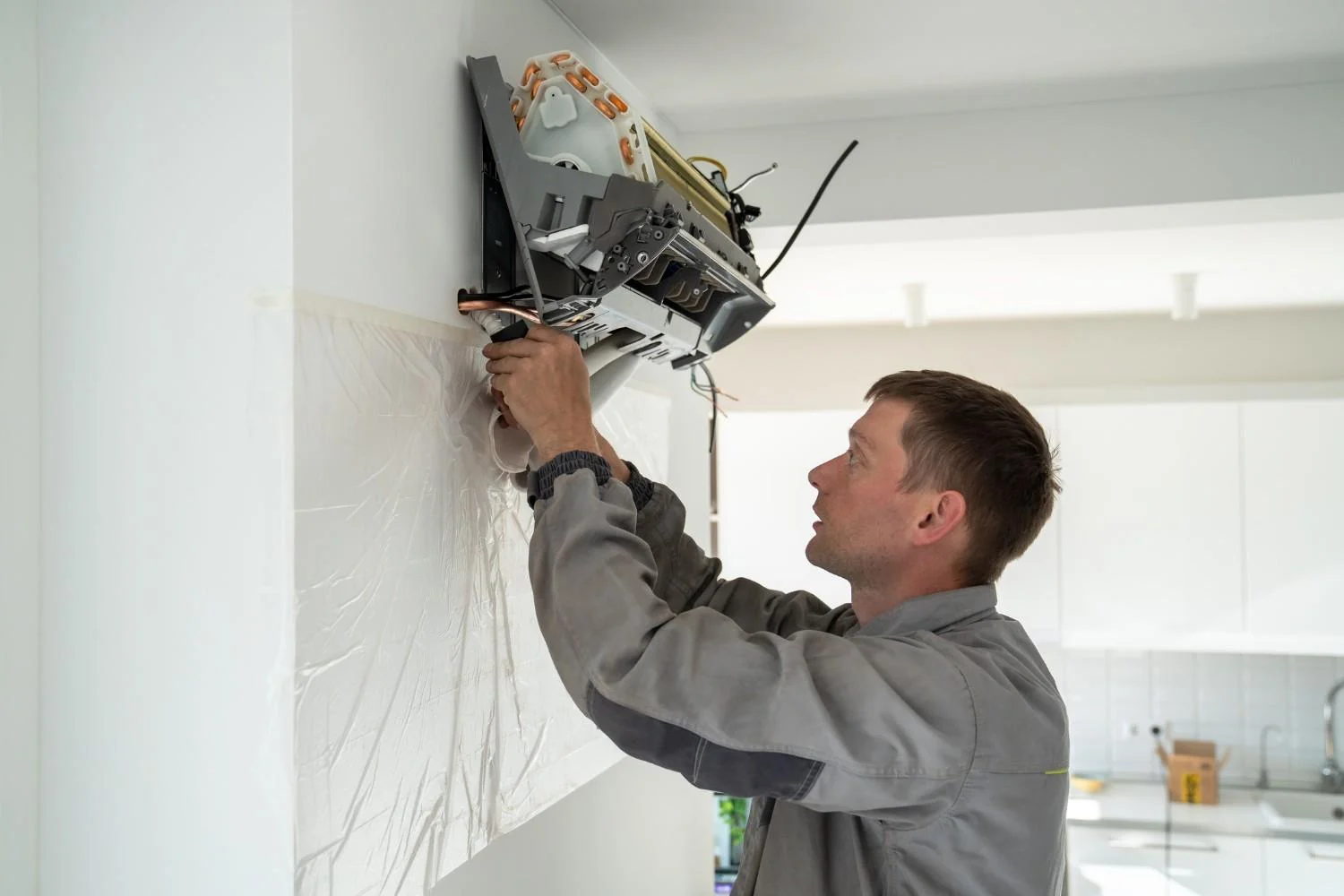 A male HVAC technician is installing a mini-split air conditioner on a white wall. He is wearing a gray uniform and is holding a component of the unit with both hands, working to connect it to the main part of the air conditioner, which is mounted on the wall. A plastic sheet is taped to the wall below the unit to protect it. In the background, there is a white kitchen with a sink.