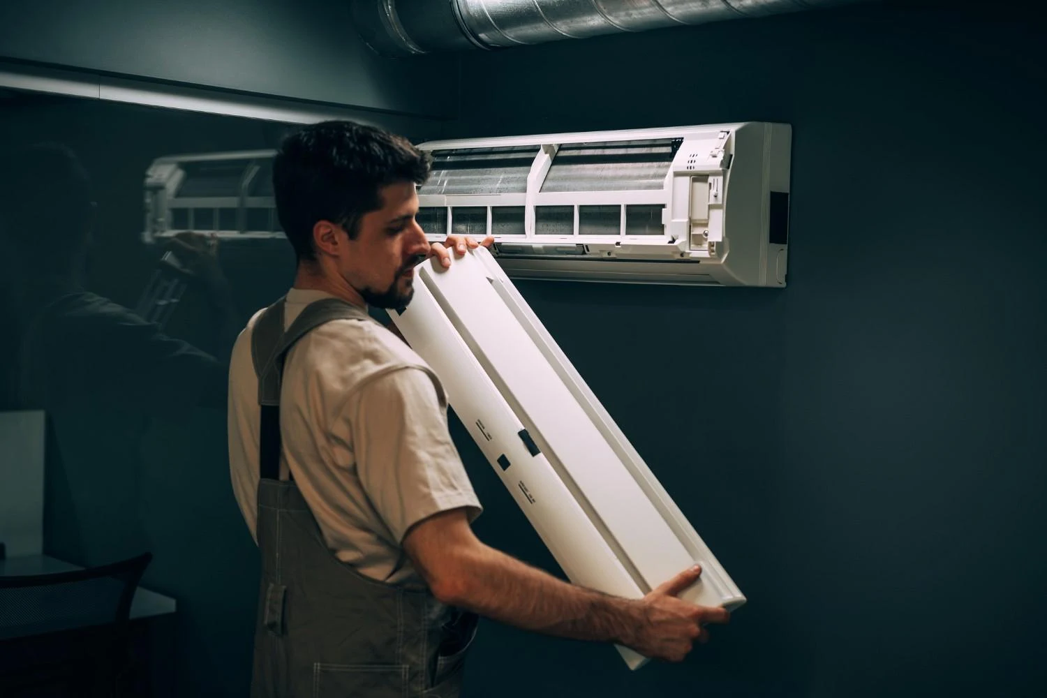 A bearded technician wearing a light-colored t-shirt and gray overalls is holding the long, white front panel of a mini-split air conditioner unit. He's preparing to attach the panel to the unit, which is already mounted on a dark gray wall. A metal vent pipe runs along the ceiling above the unit.