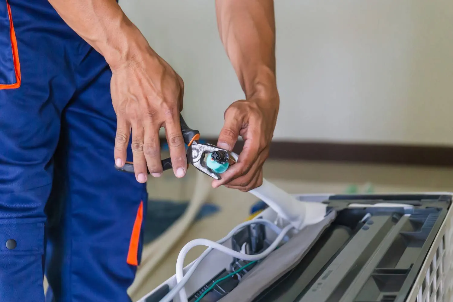 A close-up shot shows a technician's hands in blue and orange work pants. The technician is using pliers to work on a white plastic component with a blue cap, which is part of the internal wiring and tubing of a mini-split air conditioner.
