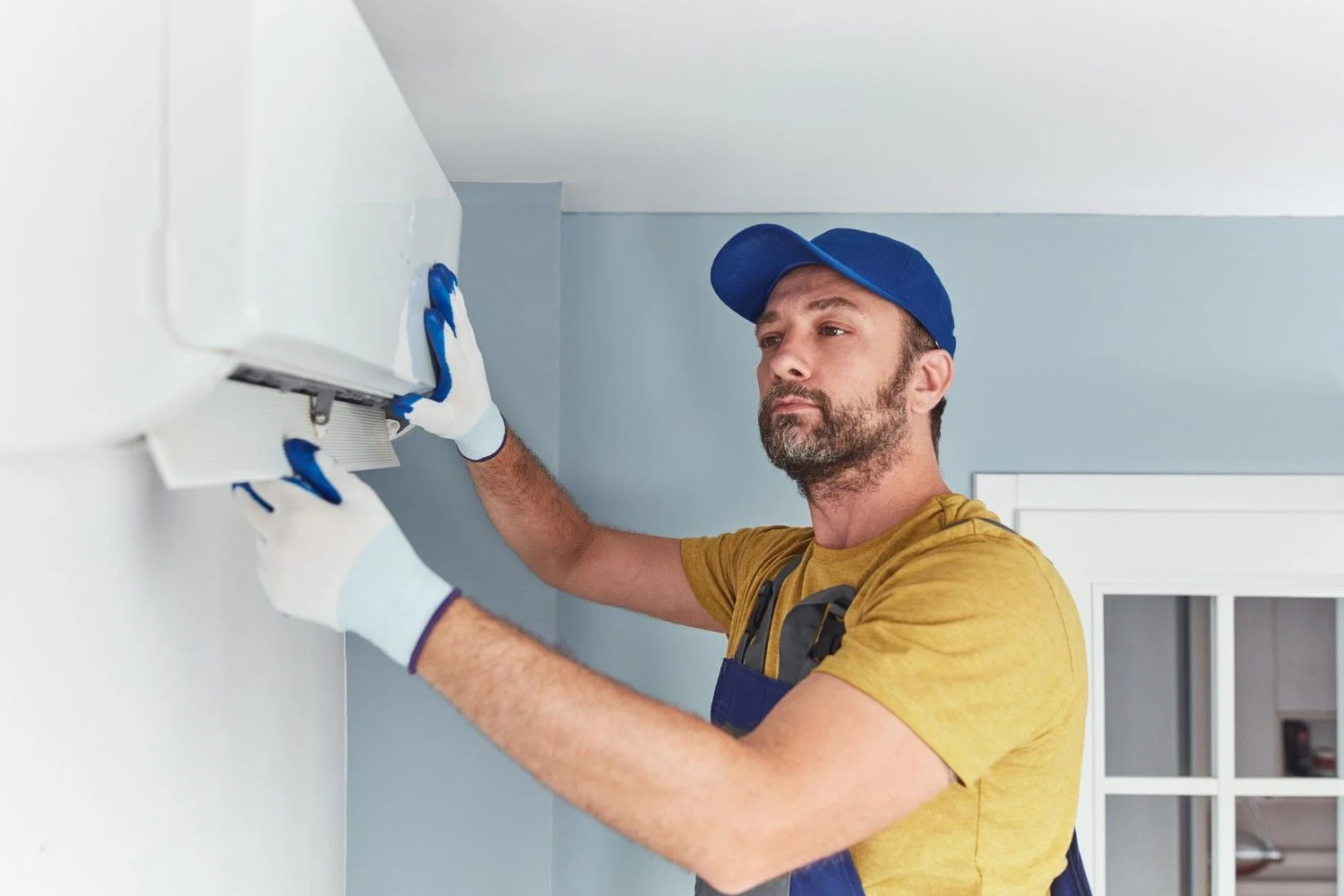 A male technician wearing a blue baseball cap, a mustard yellow t-shirt, and blue overalls is installing a white mini-split air conditioner unit. He is using both hands, covered in light blue and white gloves, to work on the unit's bottom flap or panel. The unit is mounted on a wall that is split between light blue and white colors.