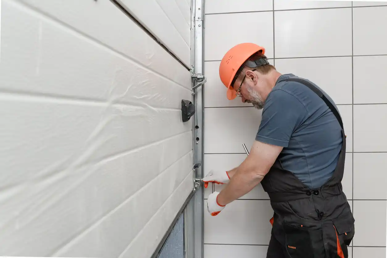 Worker in overalls and a hard hat adjusting a garage door mechanism.
