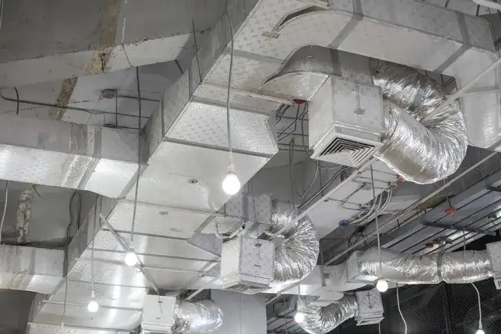 Complex arrangement of square and round silver insulated HVAC ducts and diffusers visible below a drop ceiling in a commercial space