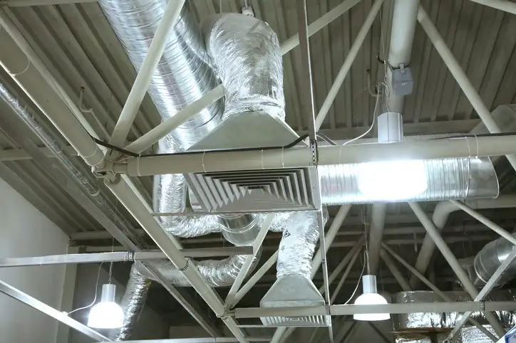 Exposed silver HVAC ductwork and diffusers mounted high in a commercial ceiling with industrial structural beams and lighting