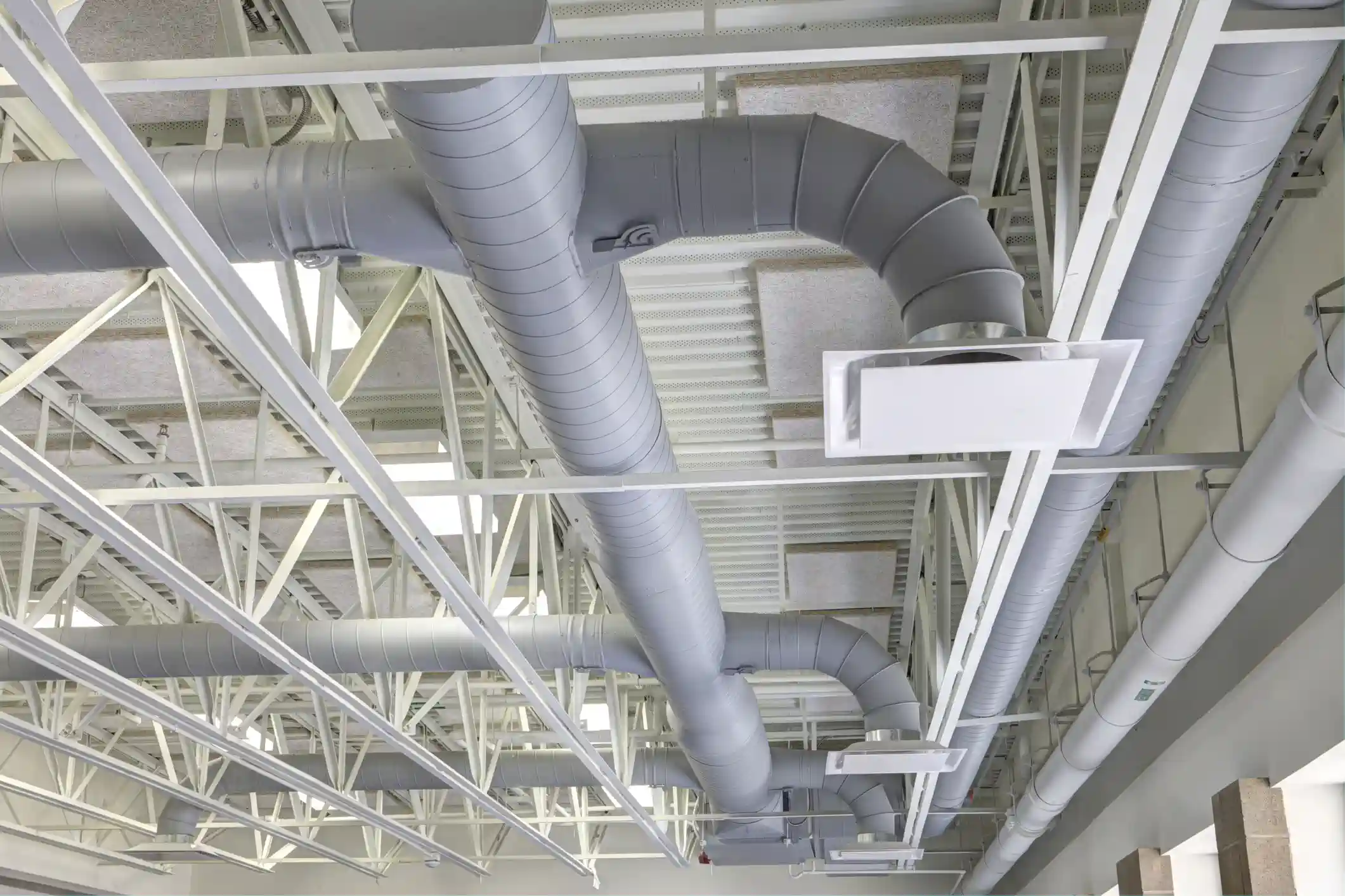 A view looking up at a high, exposed ceiling in a large commercial or industrial space. The ceiling features a network of large, cylindrical spiral metal ducts painted a uniform light gray color. The ducts run through white steel trusswork and connect to rectangular air diffusers that distribute air into the space below.