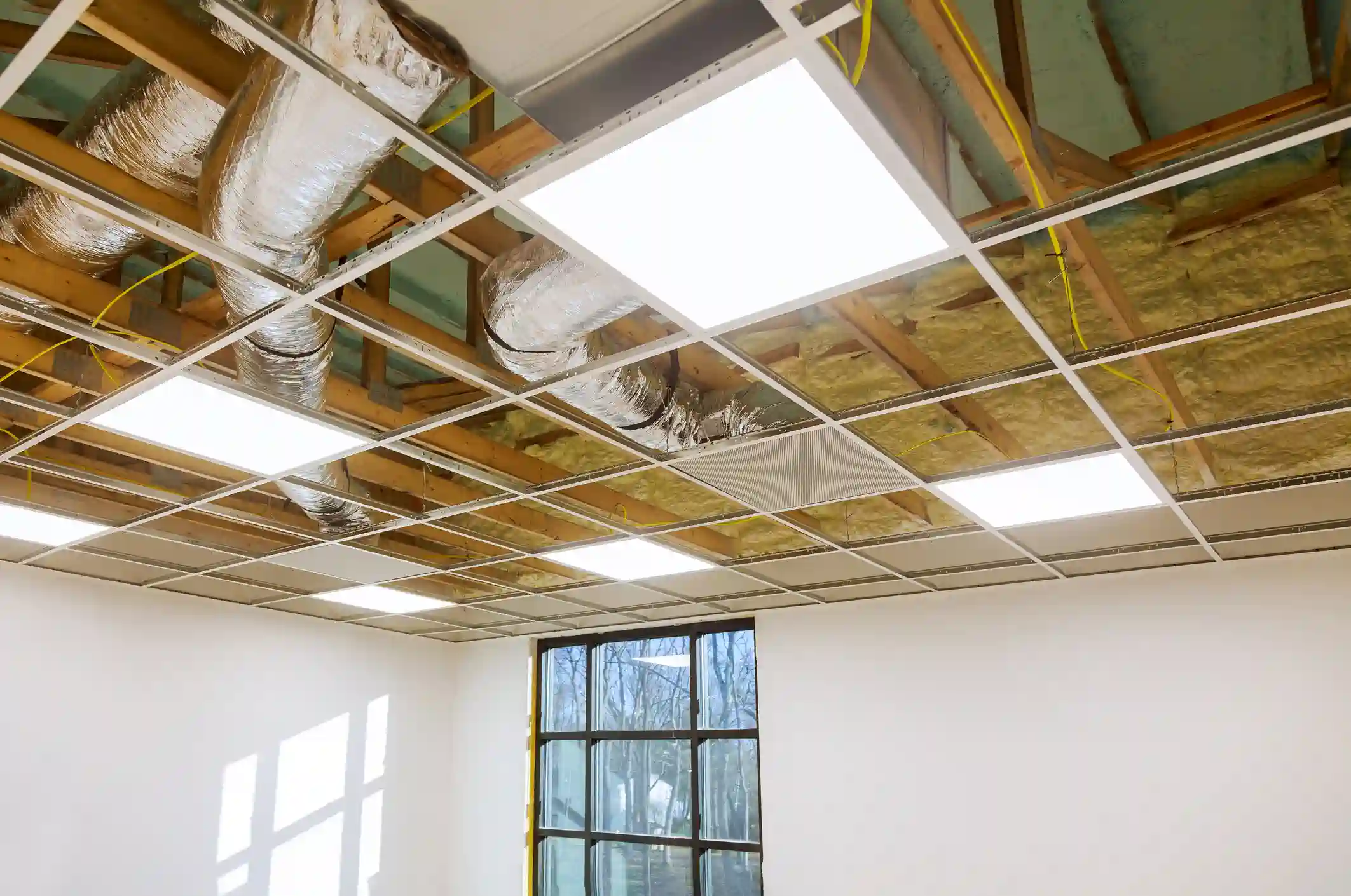 A photograph of a room during a commercial renovation, showing a drop ceiling grid system with some panels already installed as bright LED light fixtures. Above the grid, the ceiling cavity is exposed, revealing wooden joists, yellow fiberglass insulation, and runs of large, flexible, silver foil-insulated ductwork for the HVAC system.