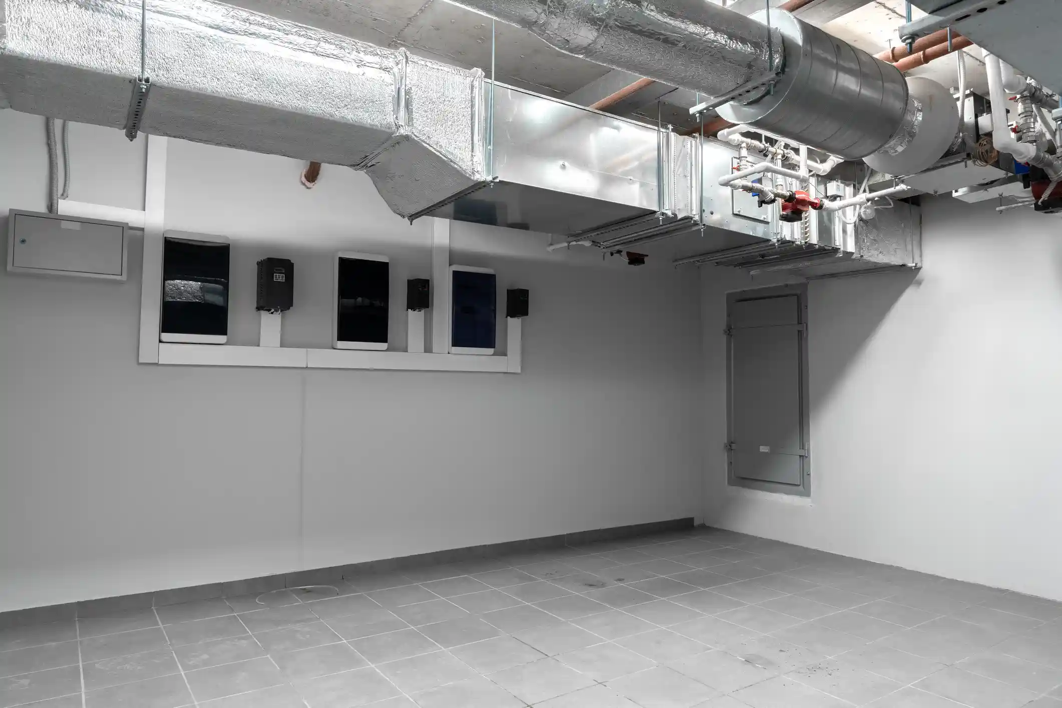 A view of an empty, light-gray room, likely a mechanical or plant room, featuring exposed HVAC ductwork and utility lines along the ceiling. A large, rectangular galvanized sheet metal duct runs high across the room, with a round, insulated metal duct and various pipes and valves visible above and to the right of it. On the back wall, several dark control panels or electrical boxes are mounted above a horizontal ledge.