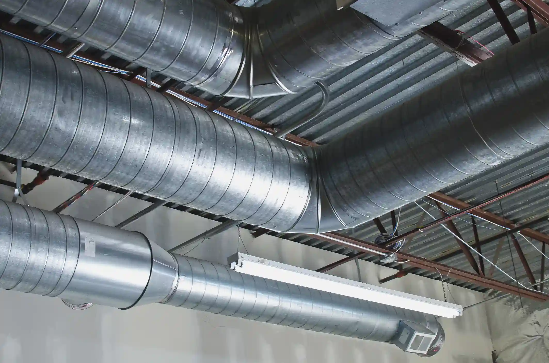 The exposed ceiling of a commercial space, dominated by a network of large, spiral galvanized sheet metal ductwork. The ducts are connected by various fittings, including a 90-degree elbow and T-junctions. A long fluorescent light fixture is mounted below one of the lower ducts, with a rectangular air diffuser visible at the end of that duct run.