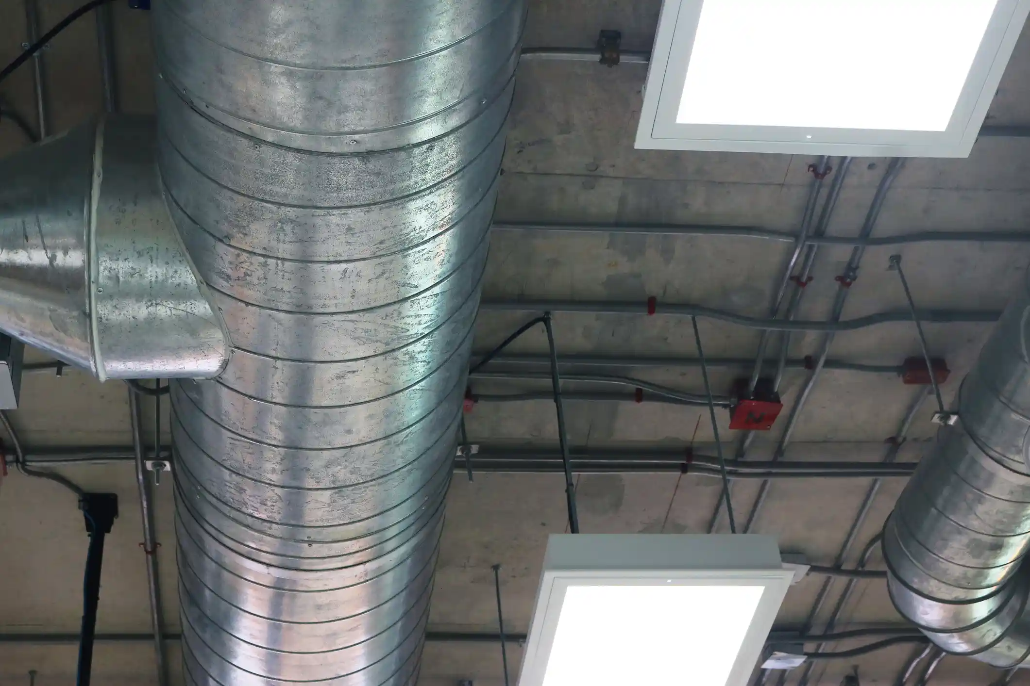 A close-up, low-angle shot of an exposed concrete ceiling in a modern industrial-style building. The image is dominated by large, unpainted spiral galvanized metal ductwork. Various electrical conduits run across the ceiling, secured by clips. Two bright, square recessed LED light fixtures are visible, contrasting sharply with the raw, mechanical aesthetic of the ceiling.