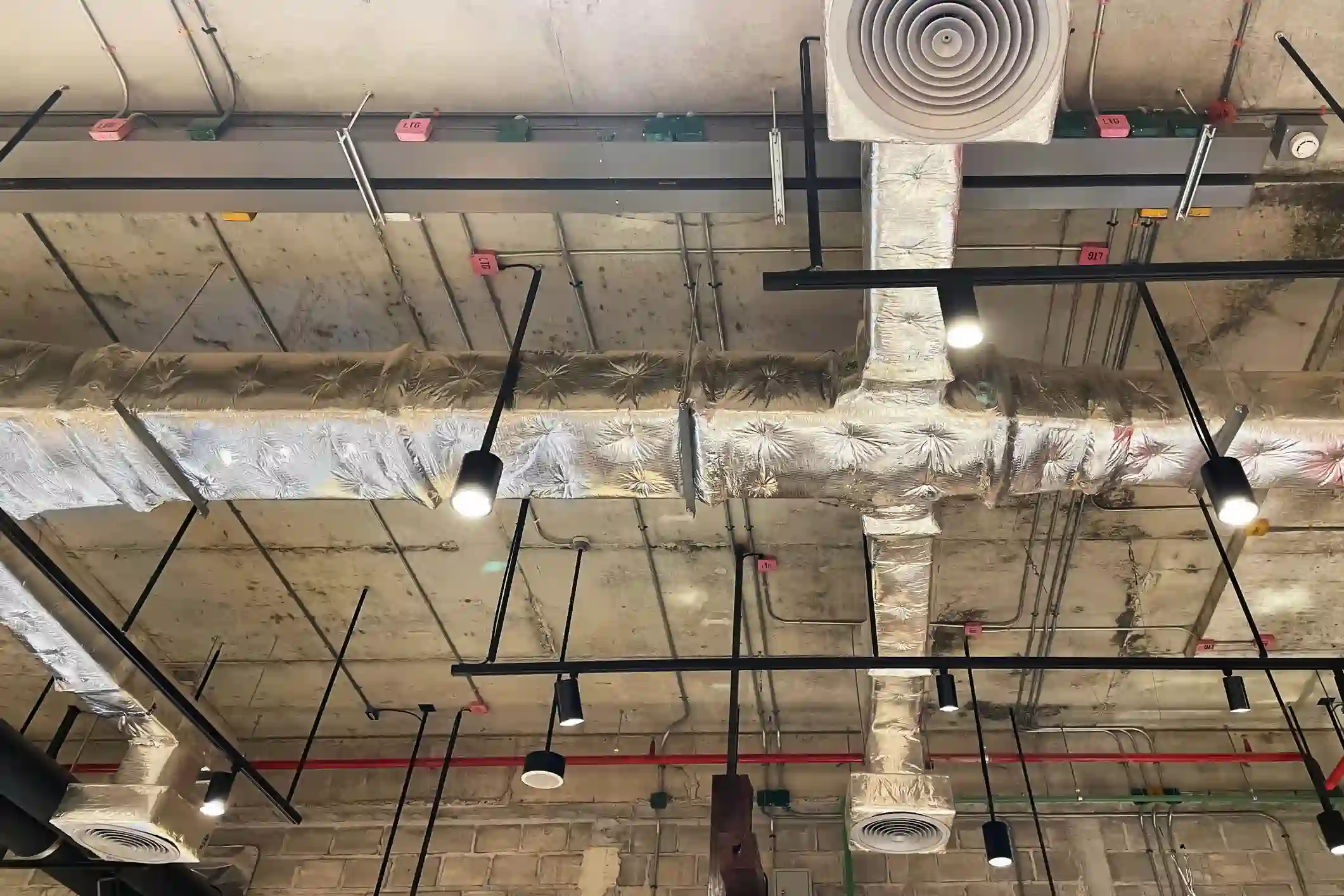 An exposed ceiling with a modern, industrial aesthetic. The ceiling features a central run of insulated HVAC ductwork wrapped in silver foil, with a round white air diffuser