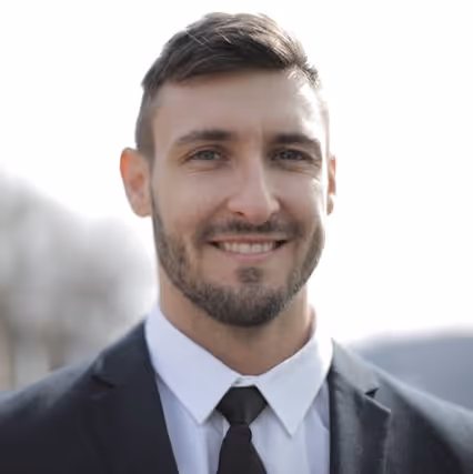 Headshot of a smiling young man wearing a dark suit and tie, standing outdoors with a softly blurred background.