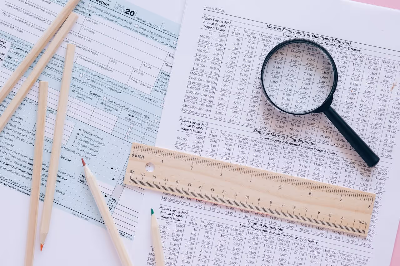 Flat lay of IRS tax forms and tables on a pink surface, with a magnifying glass highlighting numbers, a wooden ruler, and several colored pencils.