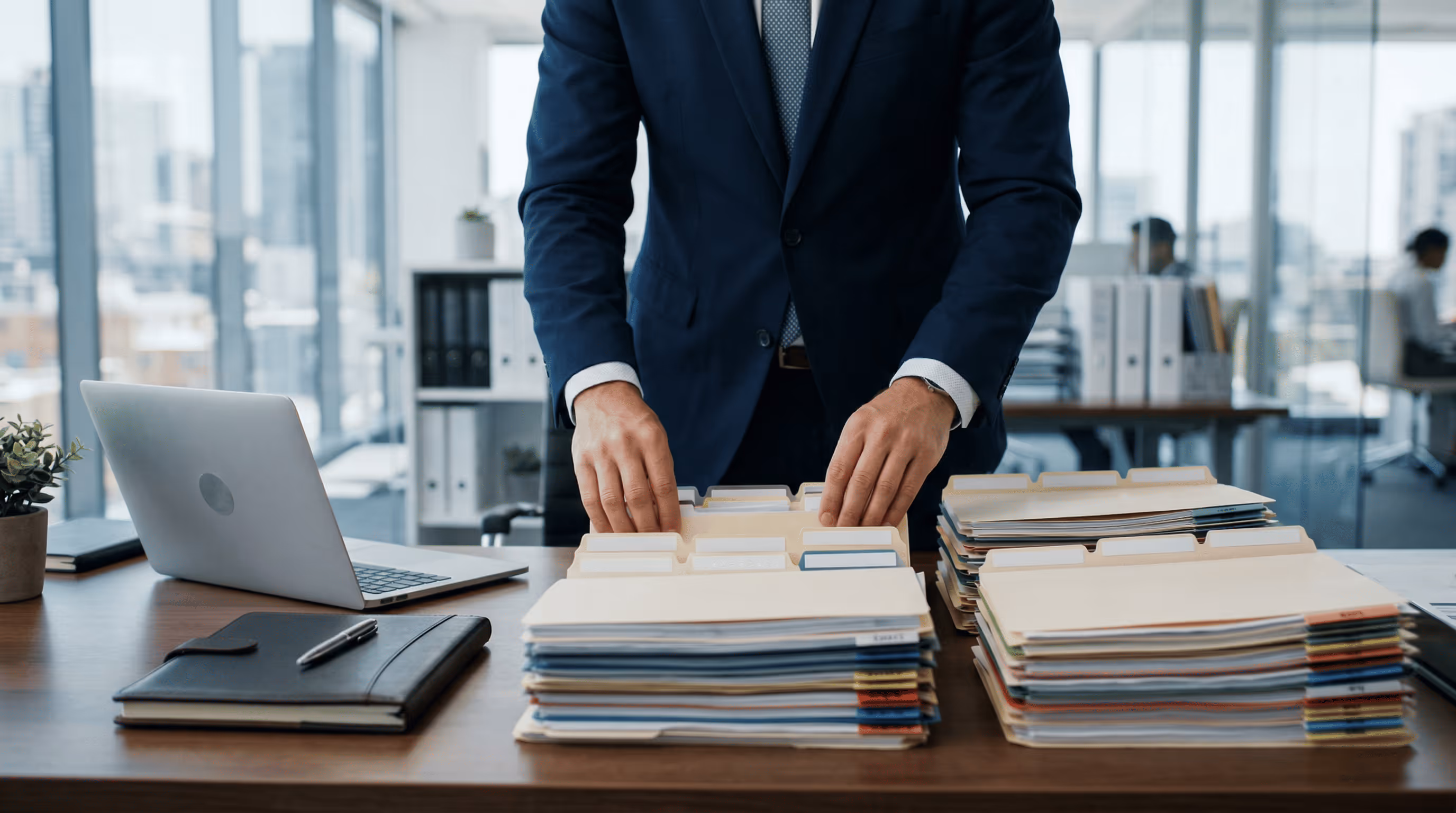 Businessman in a navy suit organizing stacks of labeled file folders on a desk with a laptop and notebook nearby in an office.