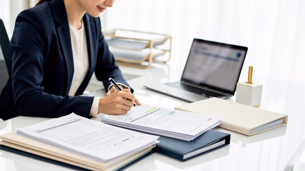 Person in business attire reviewing and marking papers at a desk with a laptop and organized folders.