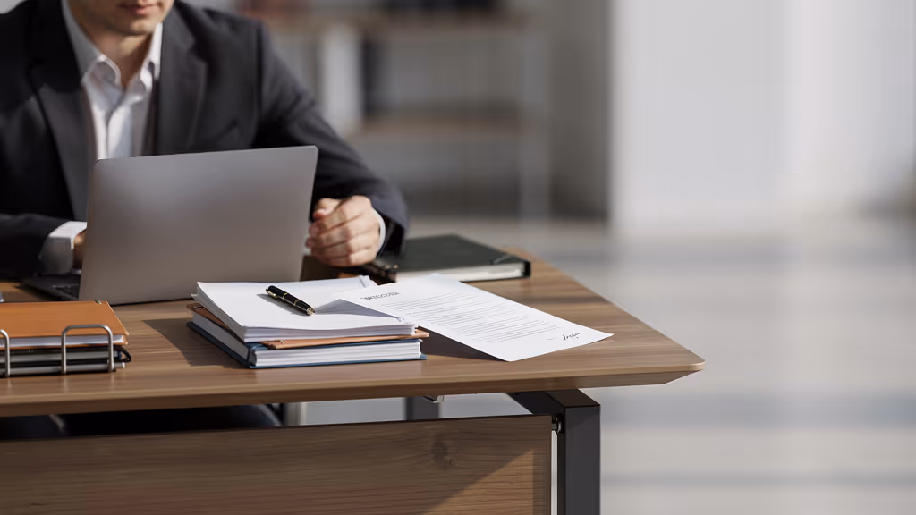Person sitting at a desk with a laptop, documents, and a pen, working in an office setting.