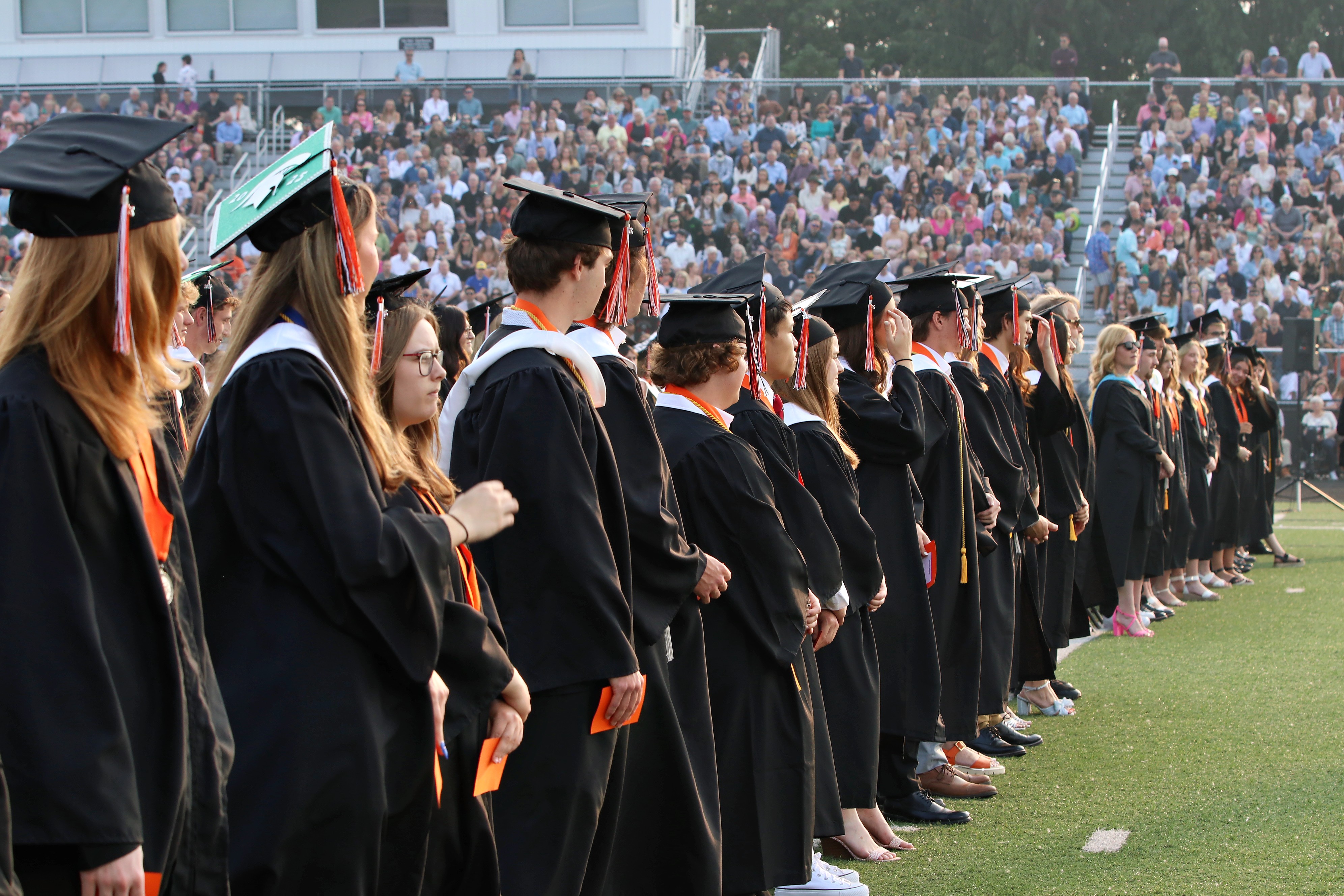 Row of graduates in black caps and gowns lined up on a field with a large audience in bleachers behind them.