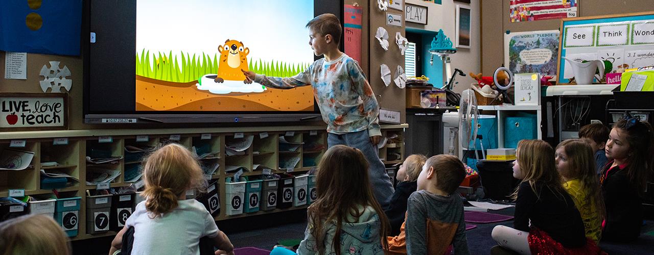 A young boy interacts with a cartoon groundhog on a large screen while other children sit on the classroom floor watching attentively.