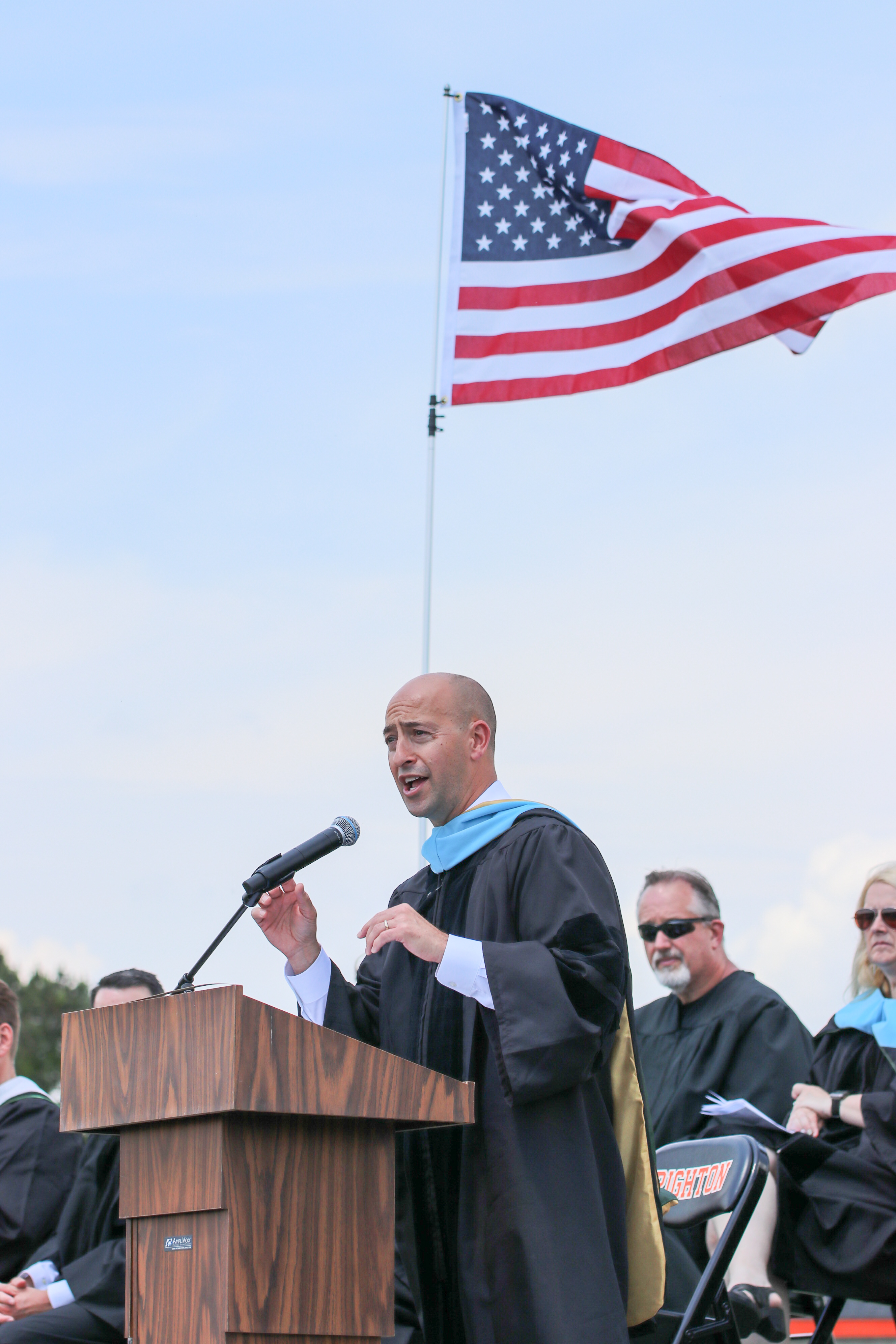 Man in academic gown speaking at a podium outdoors with an American flag in the background.