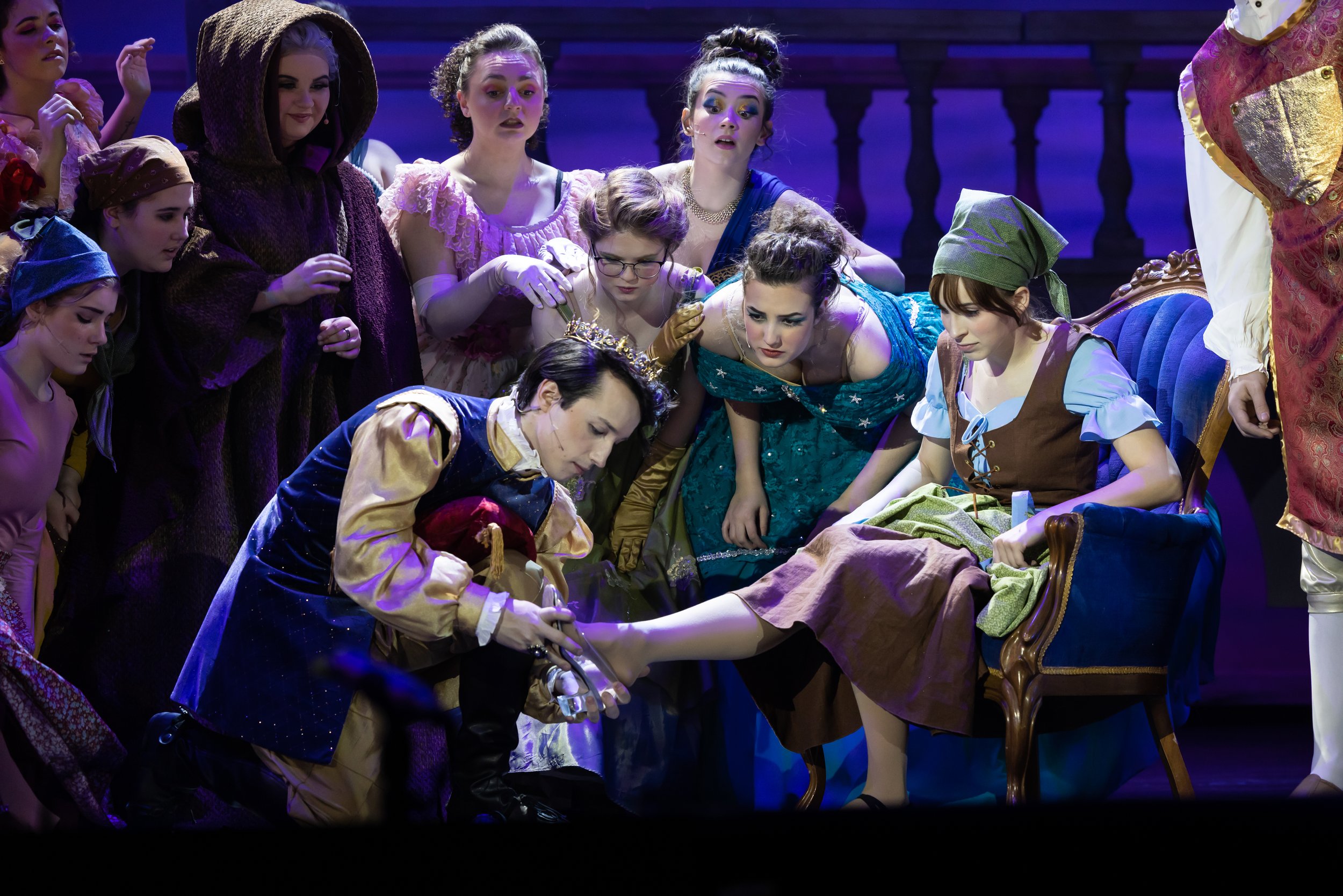 A prince kneels to fit a glass slipper on a seated woman’s foot while a group of women watch intently in a theatrical performance.