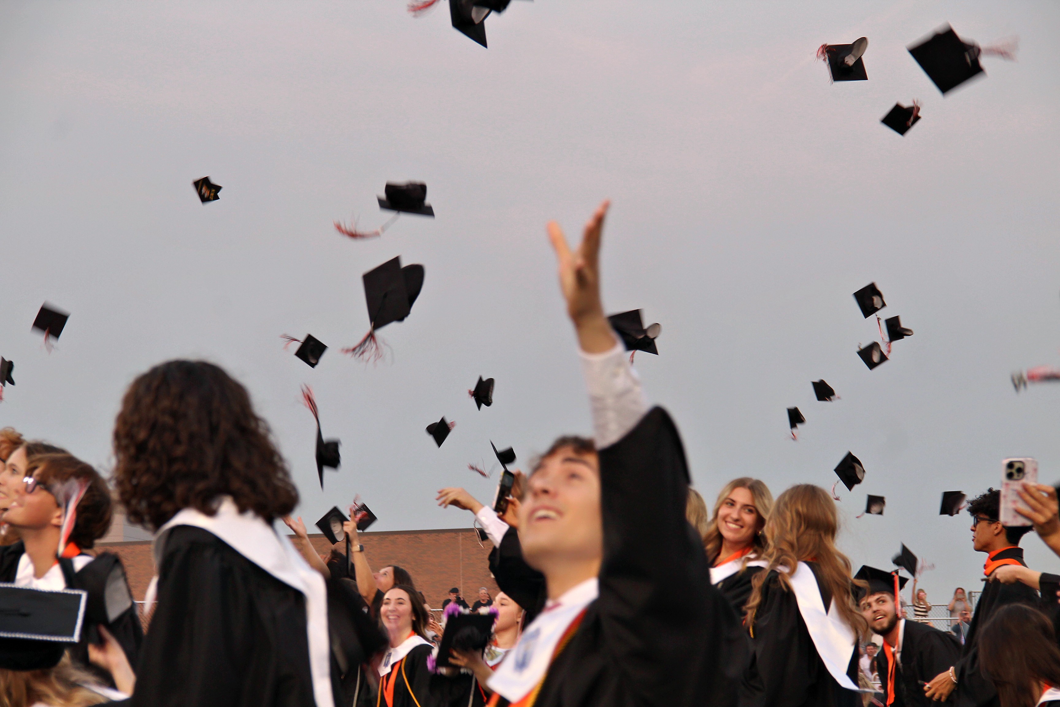 Graduates in caps and gowns celebrate by tossing their mortarboards into the air against a clear sky.