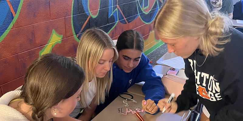 Four young women sitting and standing around a desk, focused on a craft project involving a beaded bracelet and keychain.