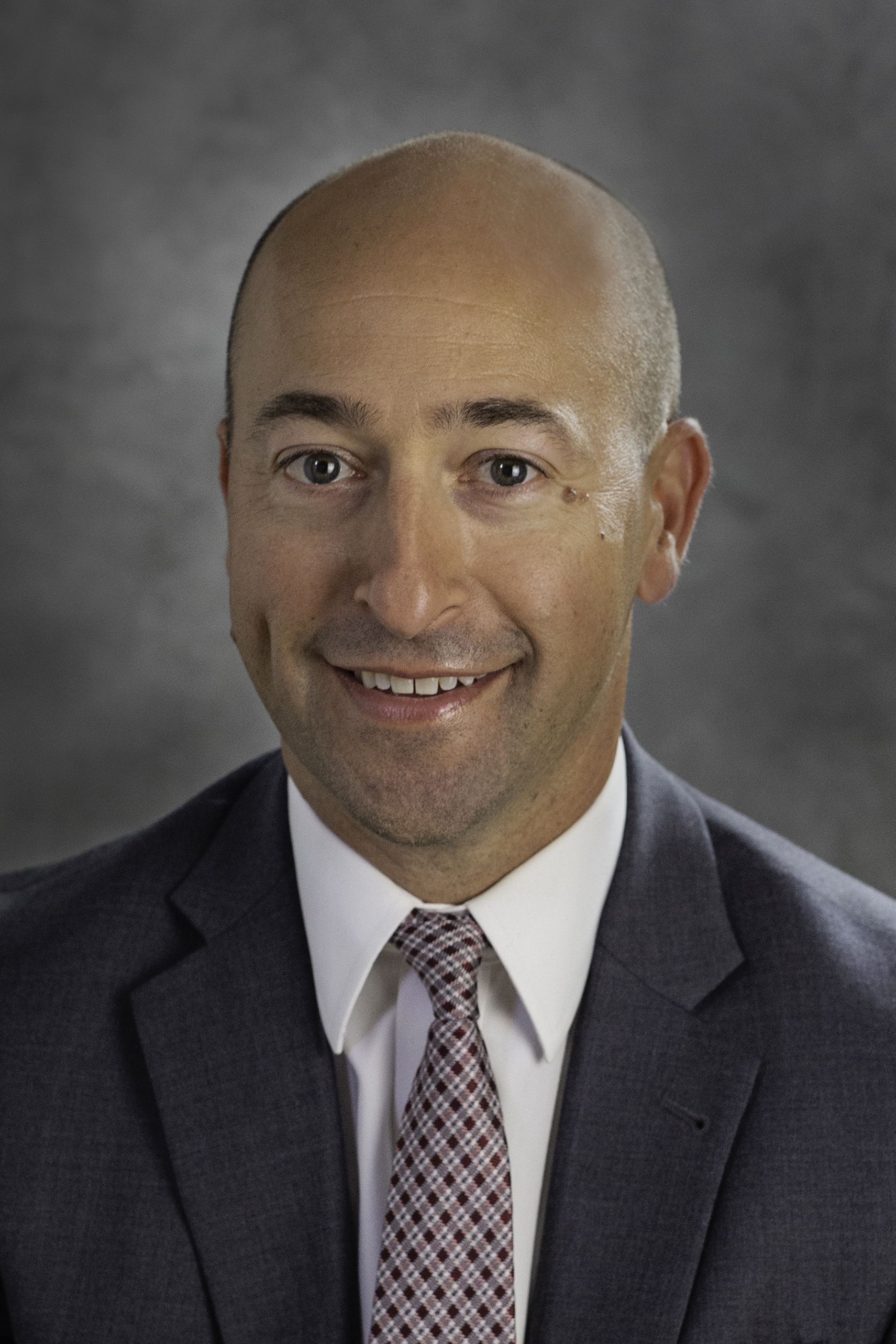 Smiling bald man in a gray suit with white shirt and patterned tie against a gray background.