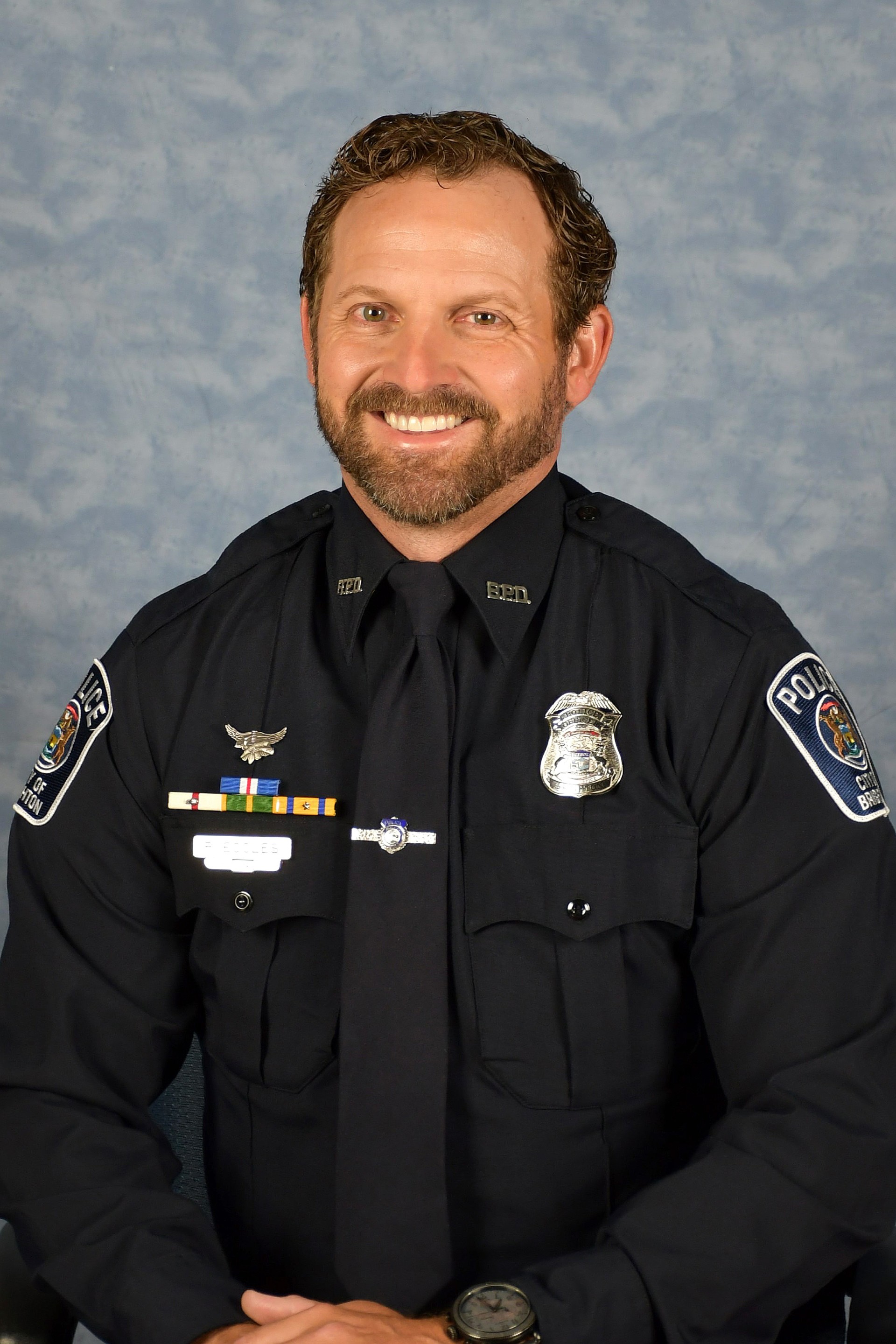 Smiling male police officer in black uniform sitting with hands folded against a gray backdrop.