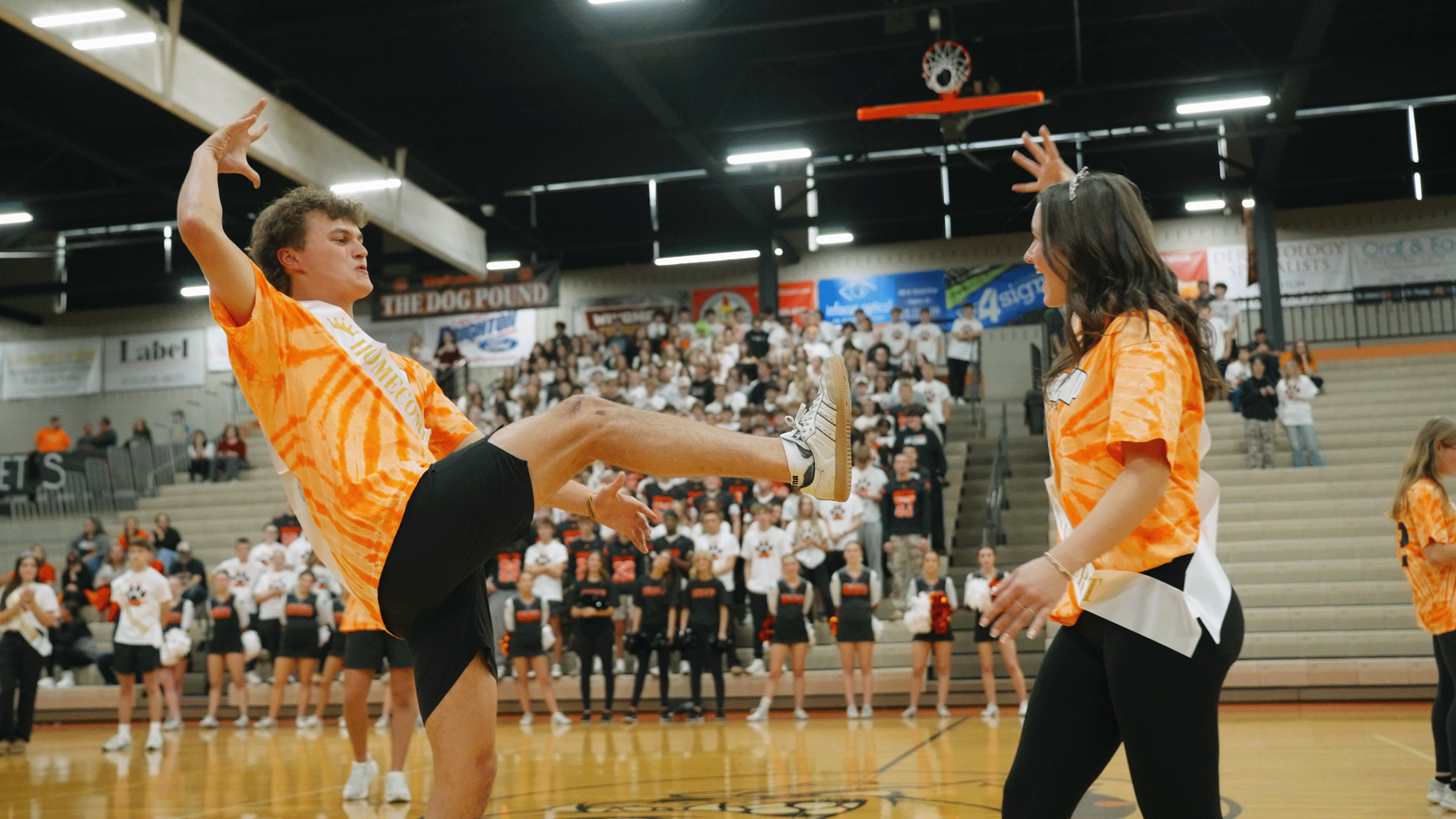 Two students wearing orange tie-dye shirts and Homecoming sashes perform a dance routine in a gymnasium with a crowd and cheerleaders in the background.