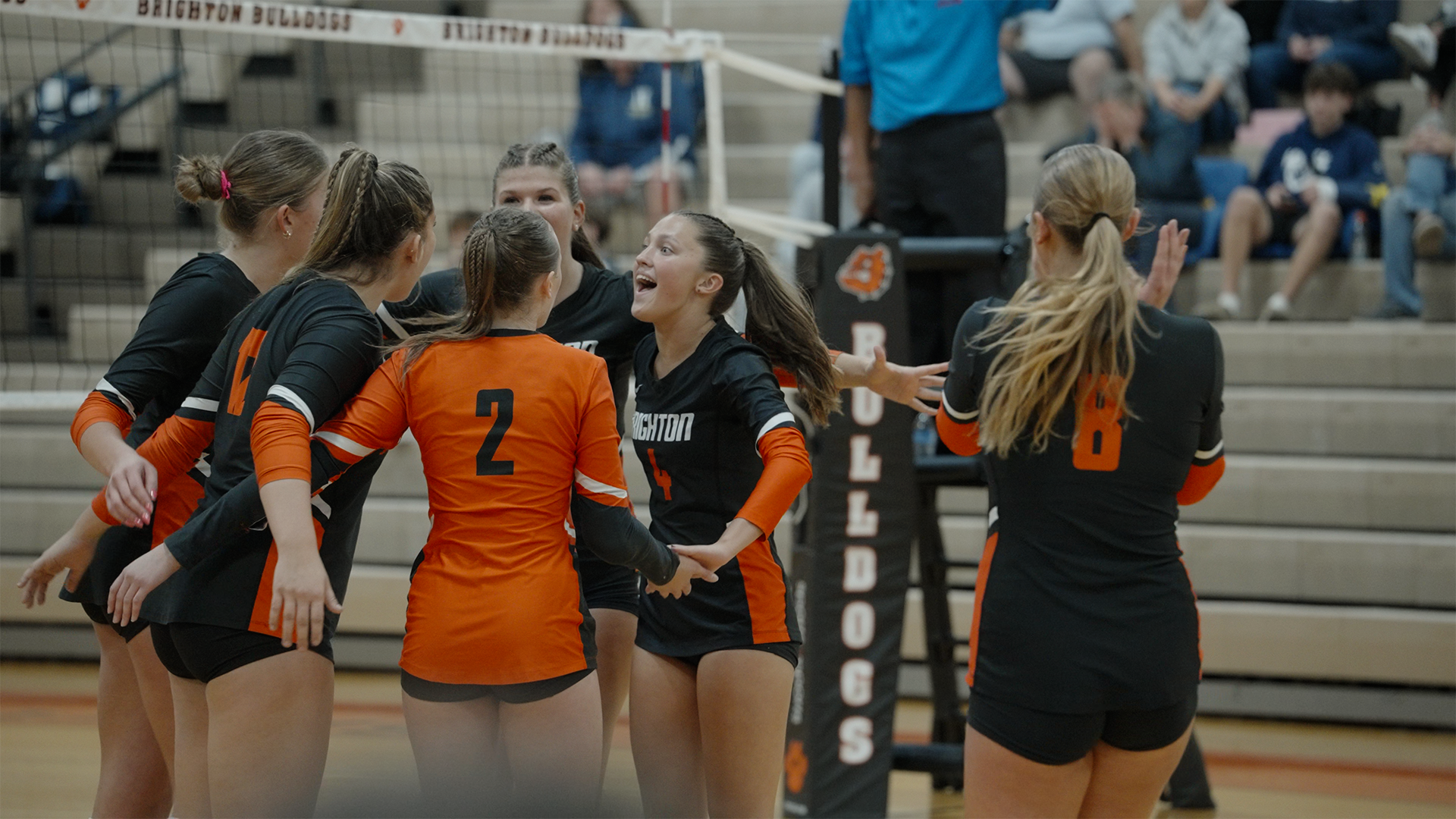 A group of female volleyball players in black and orange uniforms celebrating on the court.