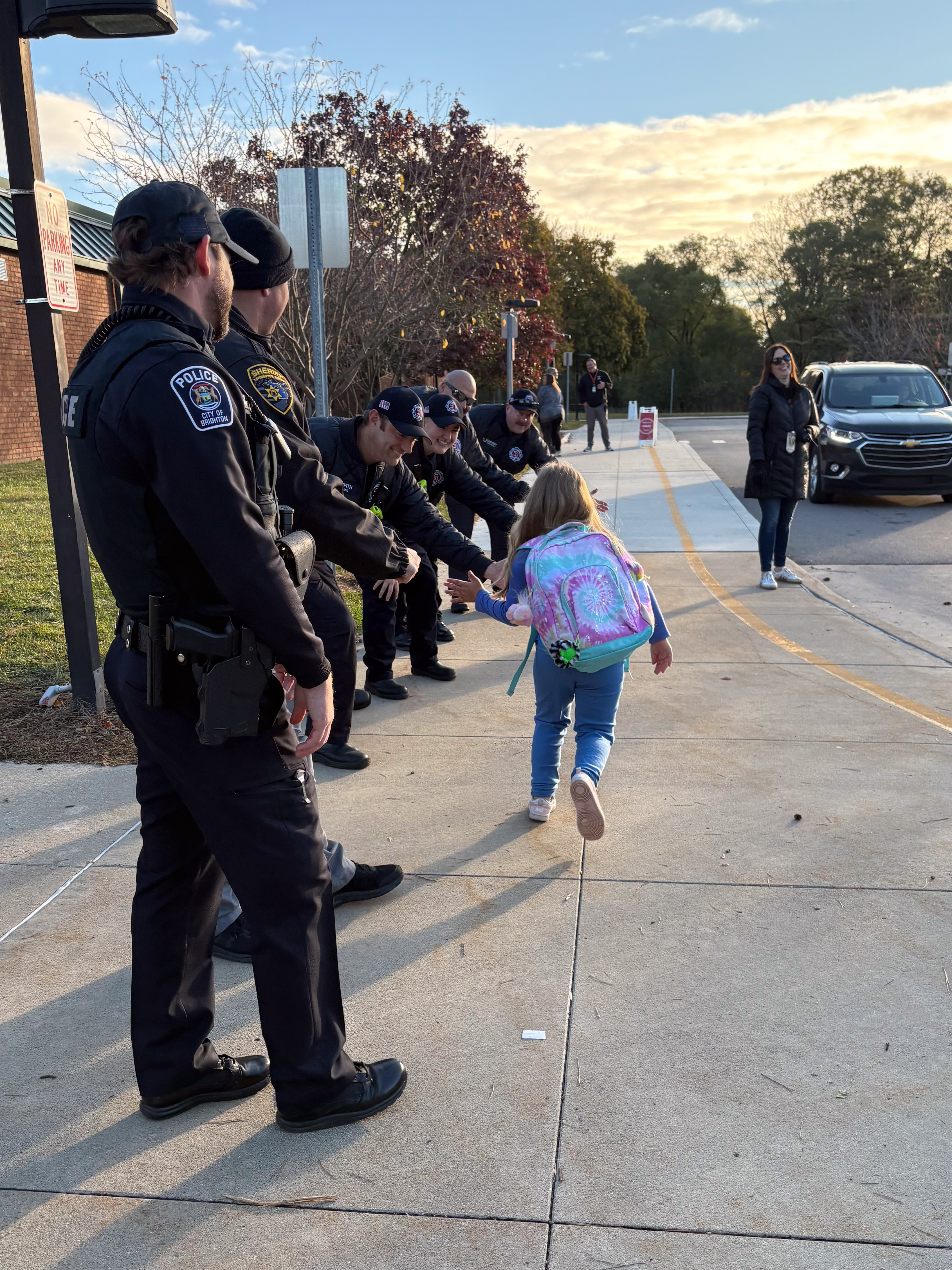 A child with a colorful backpack high-fives a line of police officers and a sheriff standing on a sidewalk at sunset.
