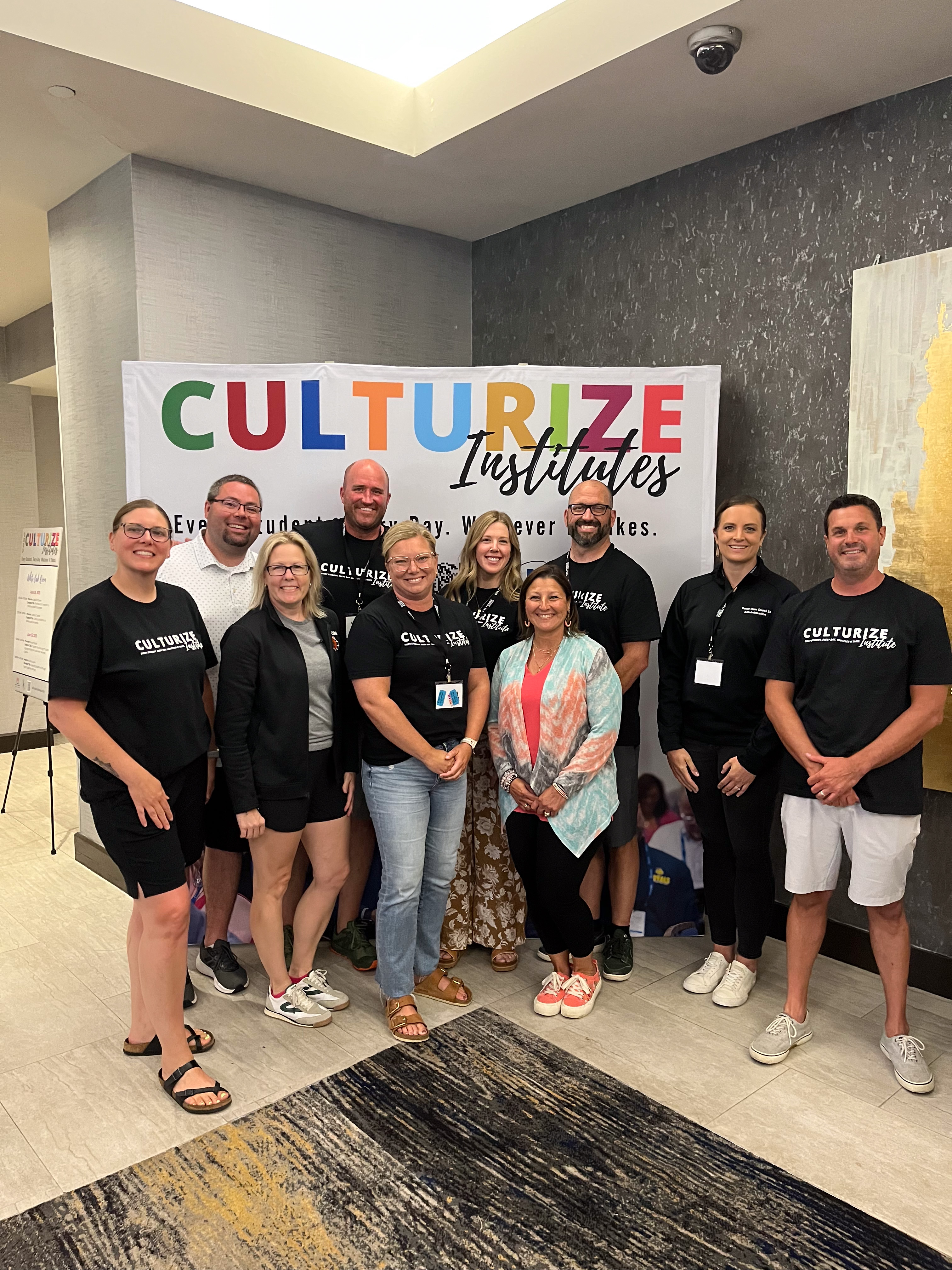 Group of ten smiling adults standing indoors in front of a colorful 'Culturize Institutes' banner.