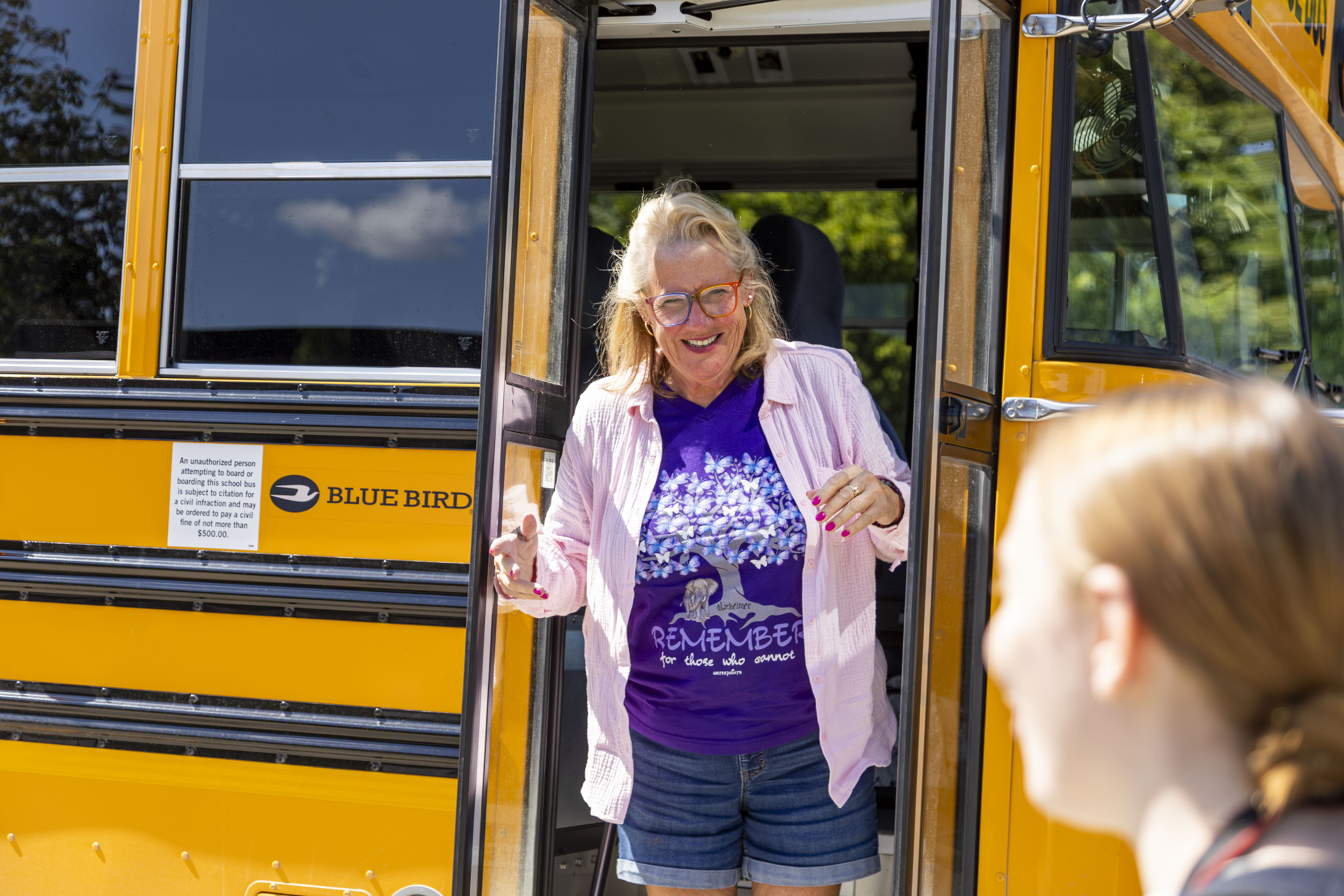 Smiling woman wearing glasses and a purple 'Remember' shirt standing in the doorway of a yellow school bus.