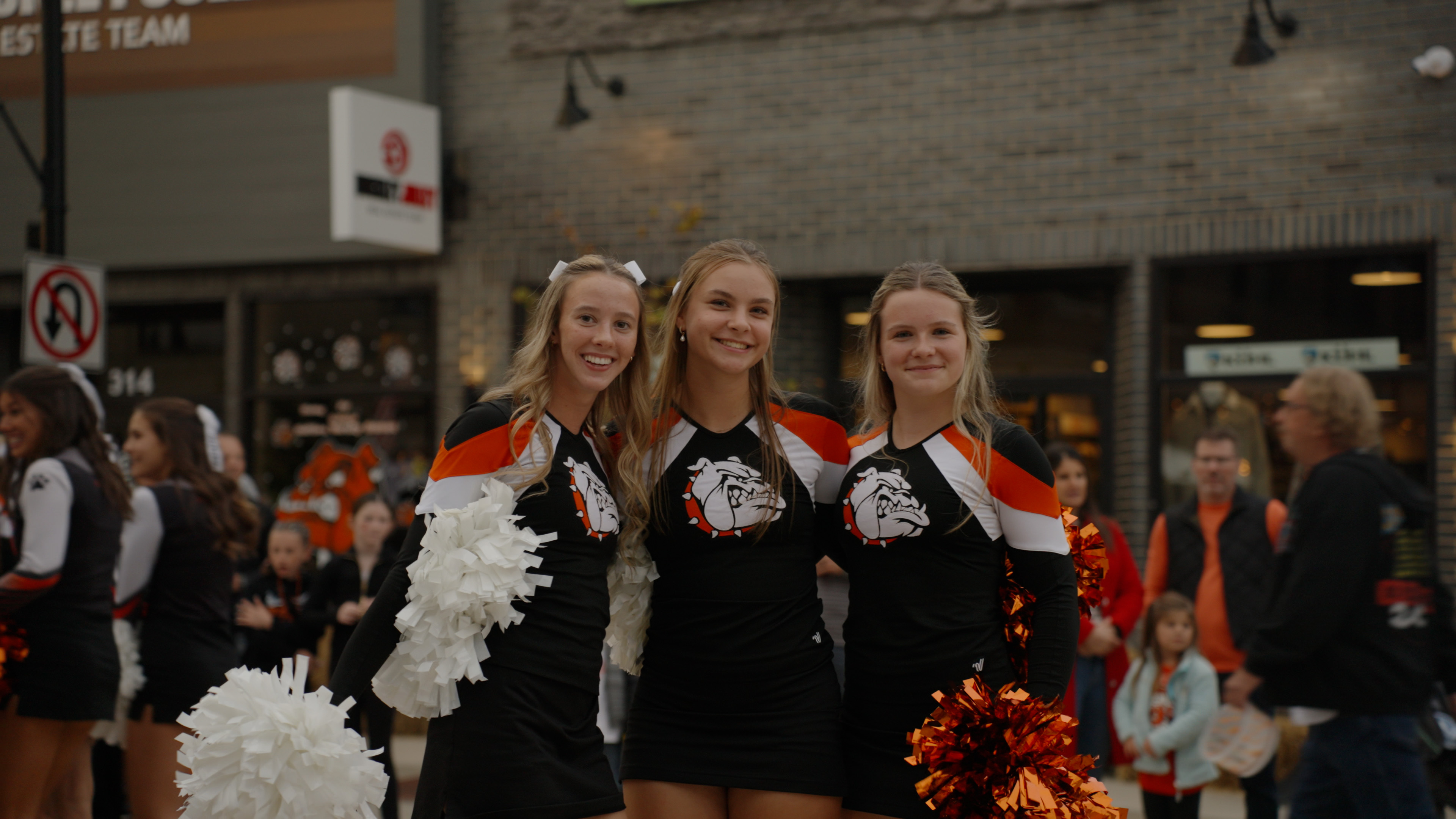 Three cheerleaders in black, orange, and white uniforms with bulldog logos smiling and holding pom-poms at an outdoor event.