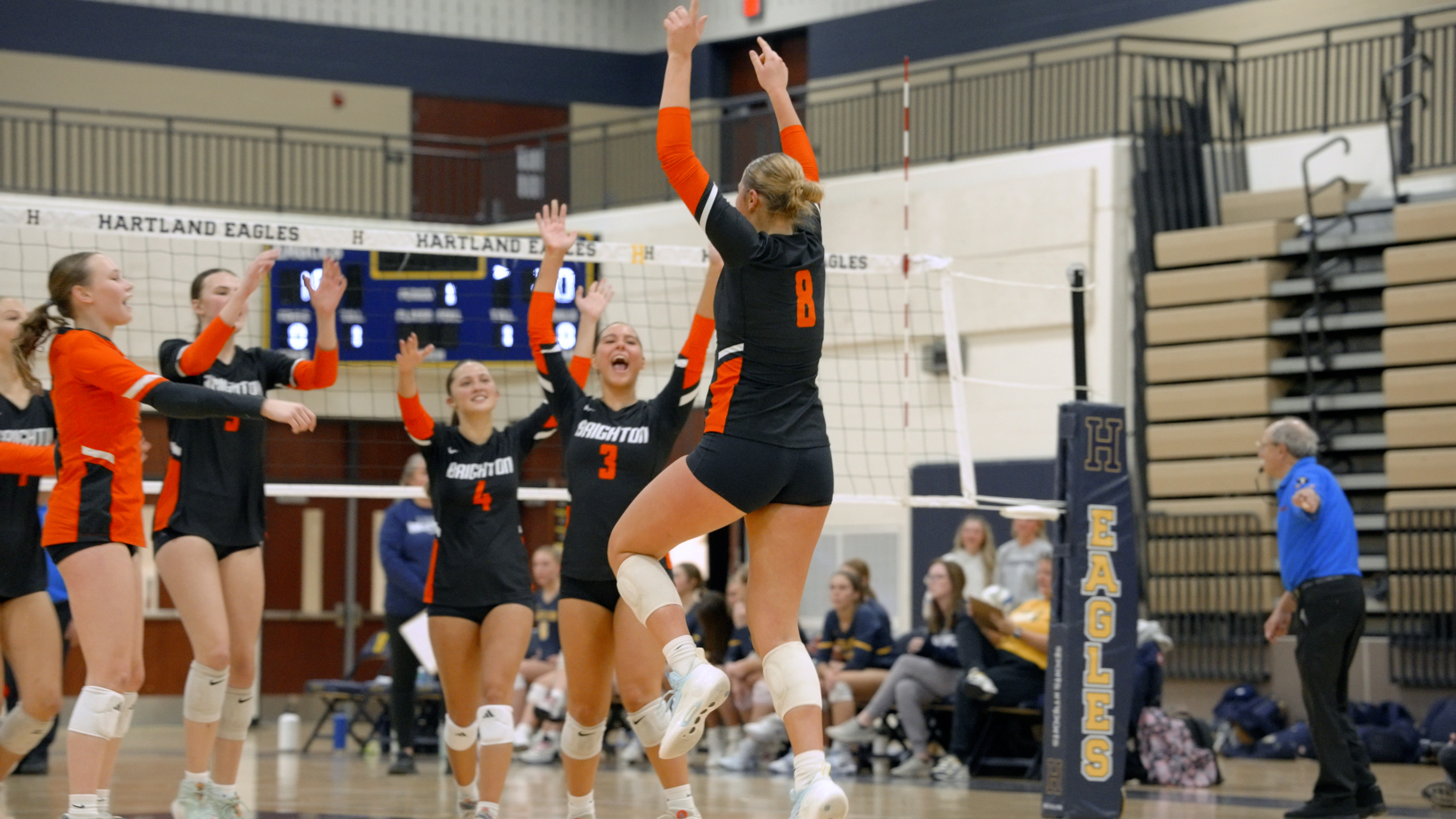 Volleyball players in black and orange uniforms celebrating on court near net in gym labeled Hartland Eagles.