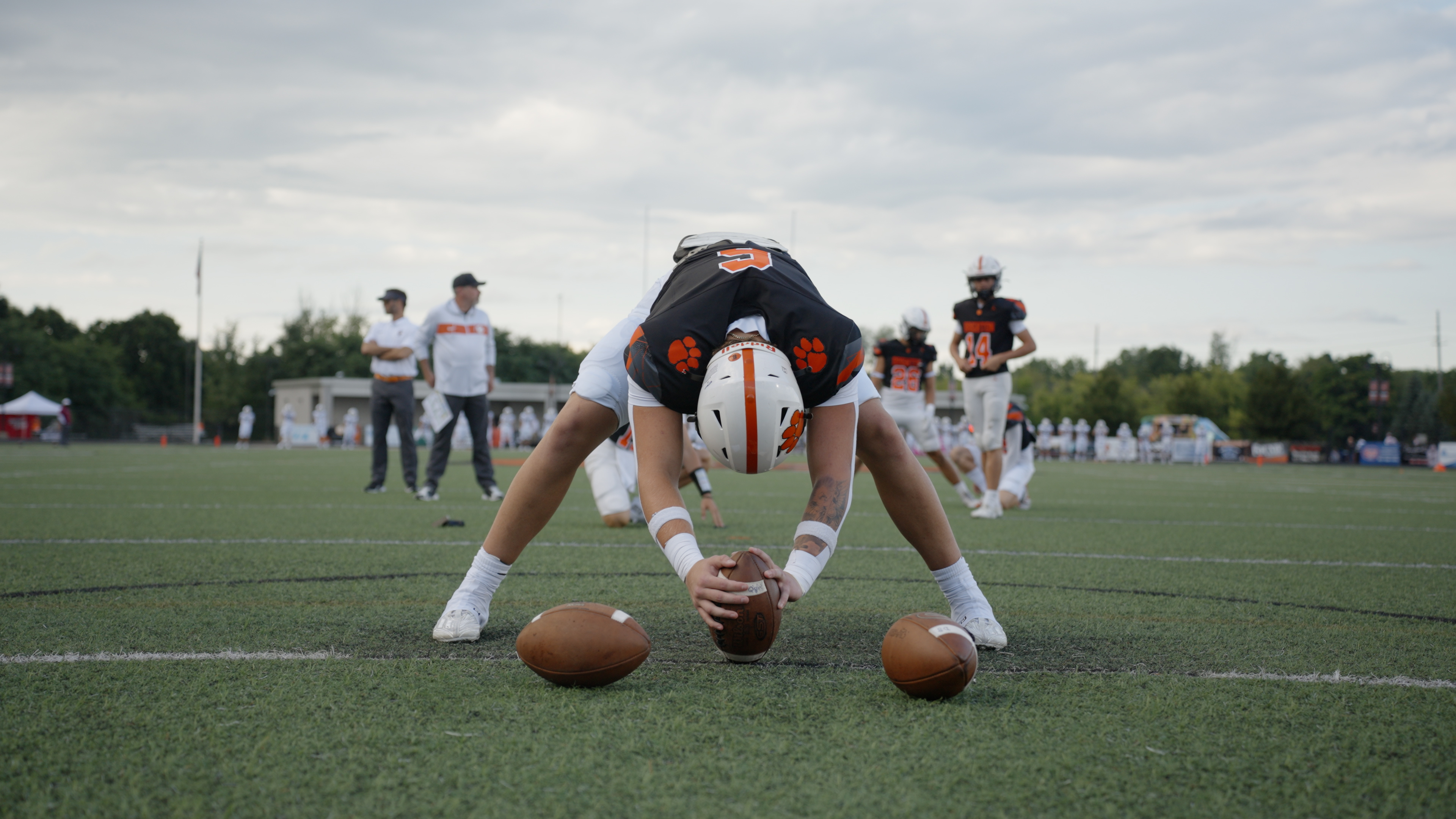 Football player in black and white uniform bending to place a football on the field with two other footballs nearby.