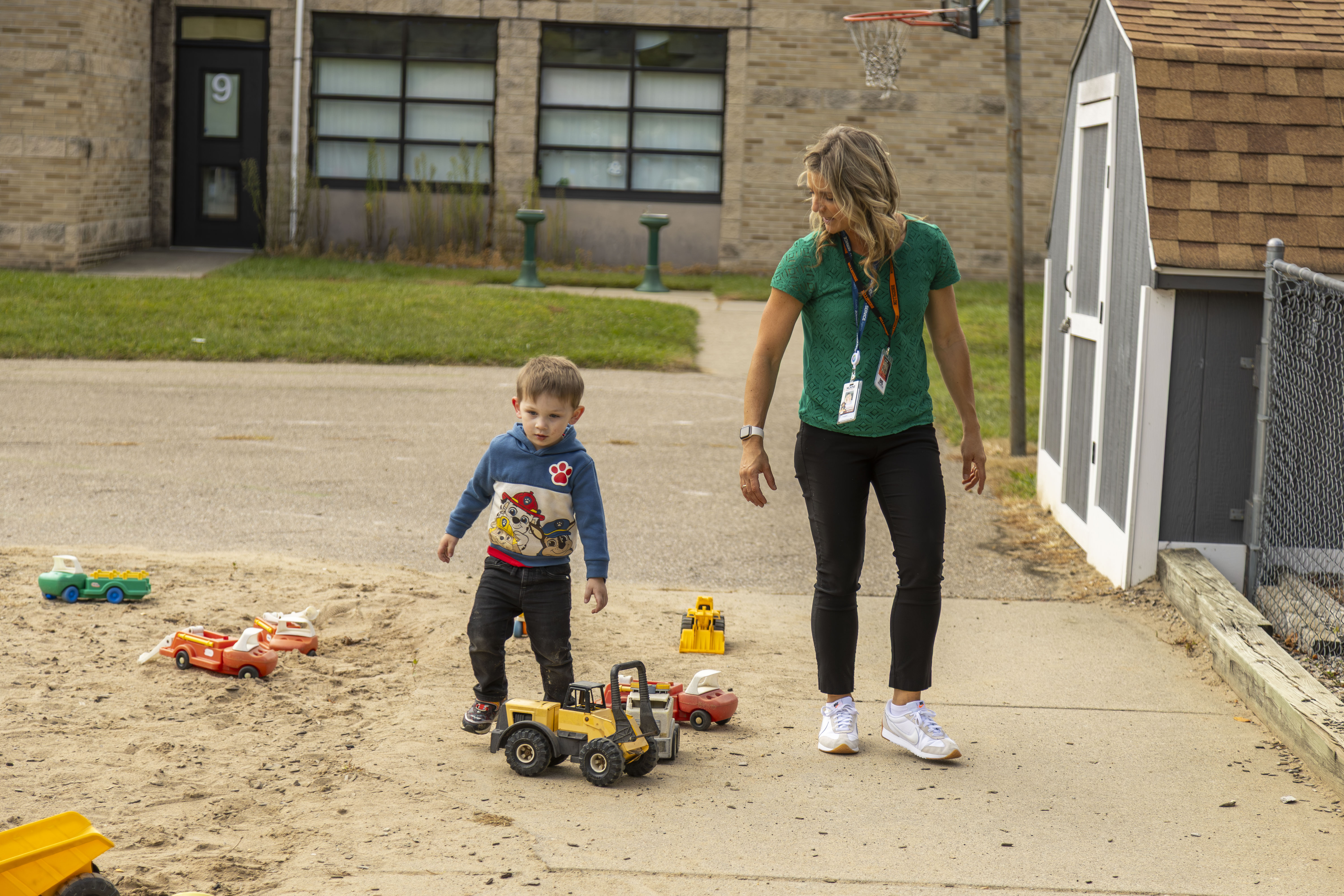 Young boy playing with toy trucks in a sandy area while a woman in a green shirt and black pants watches nearby outdoors.