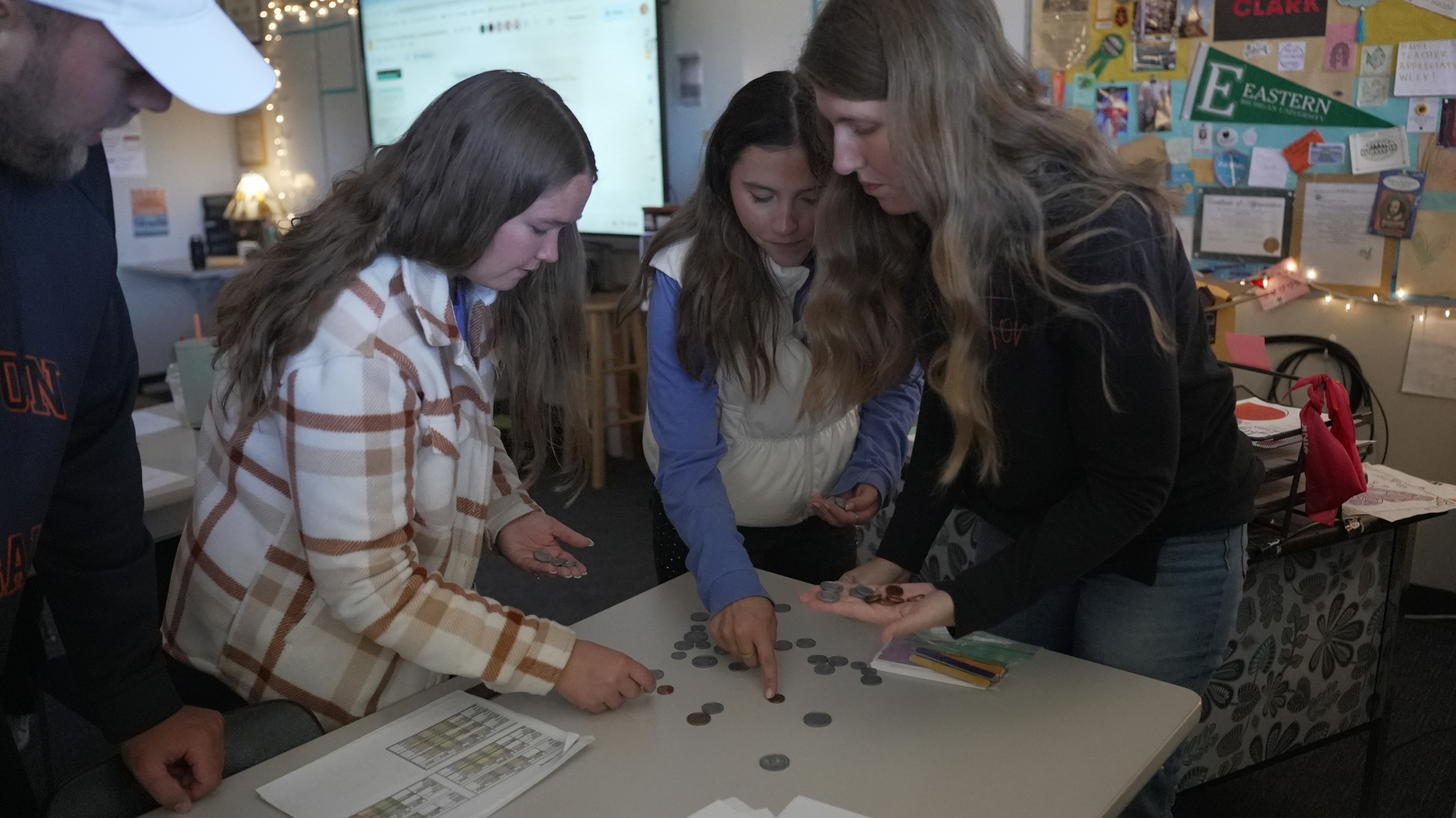Four people standing around a table counting and sorting coins in a classroom setting.
