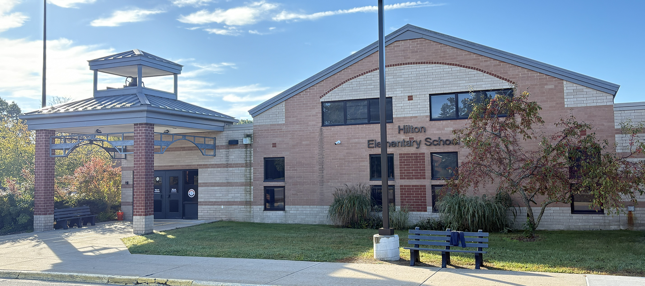 Exterior of Hilton Elementary School building with brick facade, entrance canopy, and benches on a sunny day.