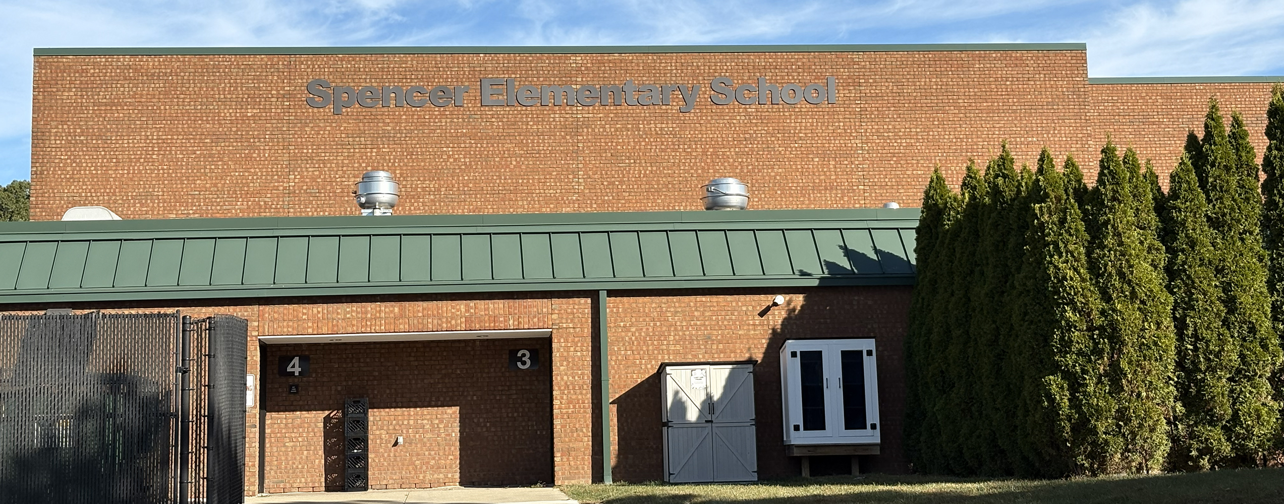 Brick building of Spencer Elementary School with green roof and tall evergreen trees on the right.