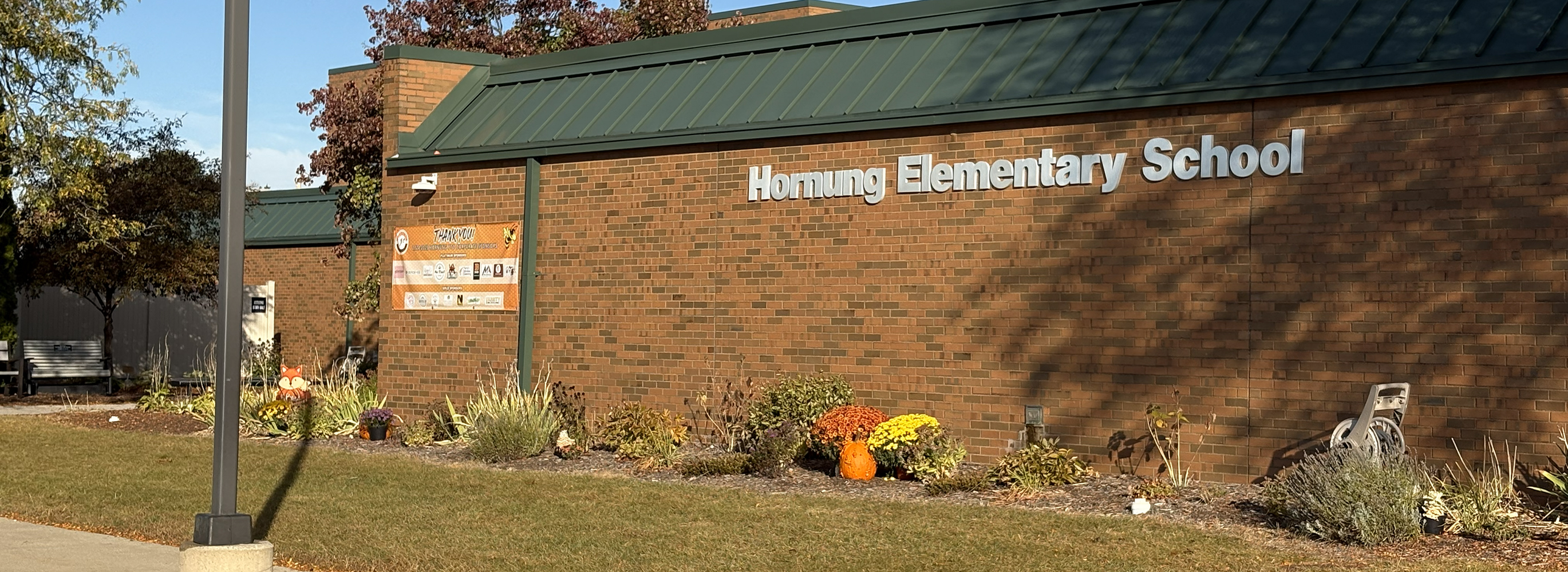 Brick exterior wall of Hornung Elementary School with a green metal roof, a garden bed with flowers and a pumpkin, and a lamp post in the foreground.