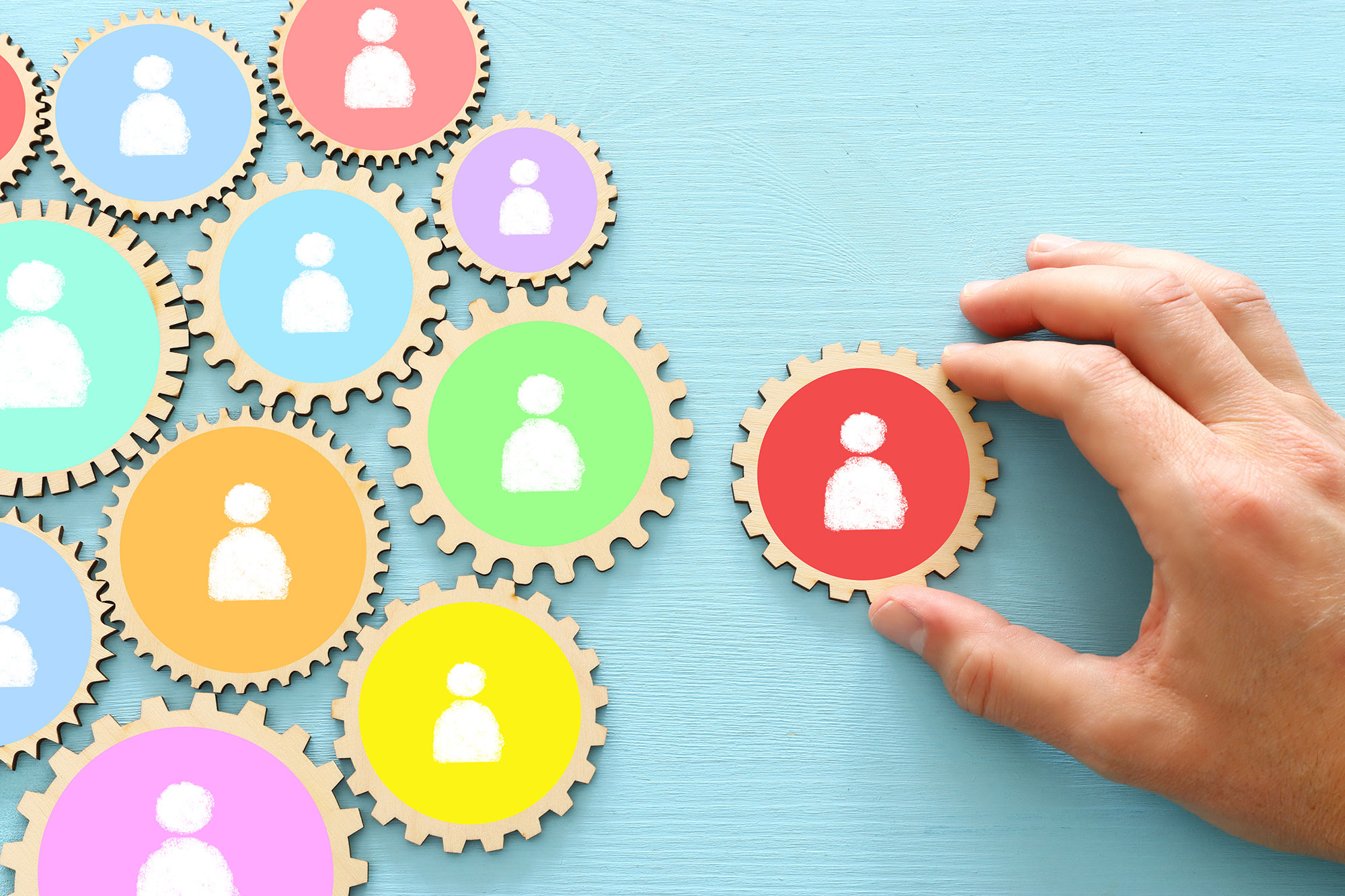 Hand holding a red wooden gear with a white person icon, near several interlocking colorful gears with similar icons on a blue wooden surface.