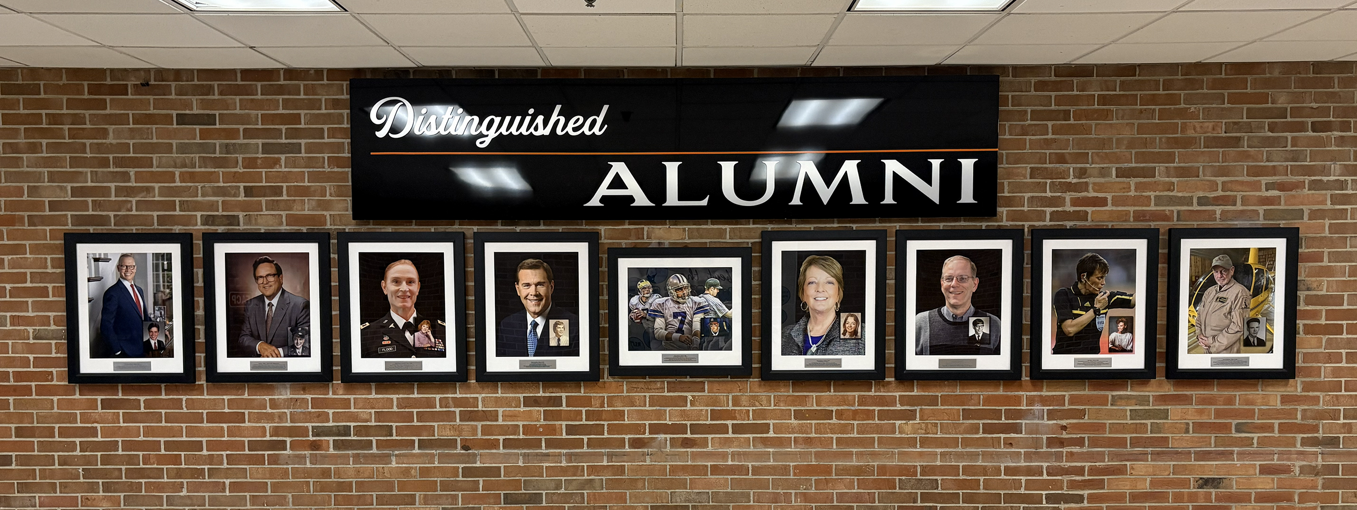 Wall display with a black sign reading 'Distinguished Alumni' above eight framed portraits of alumni on a brick wall.