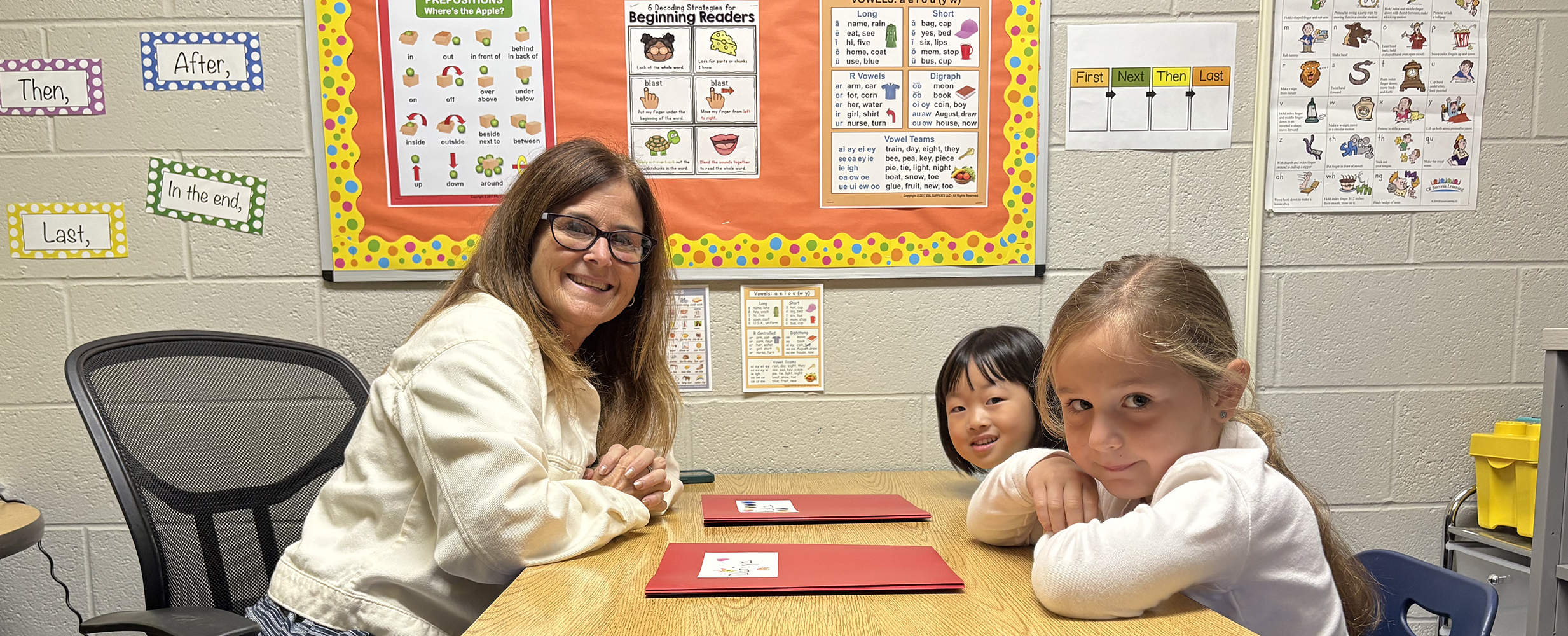Teacher smiling and sitting at a table with two young students in a classroom with educational posters on the wall.