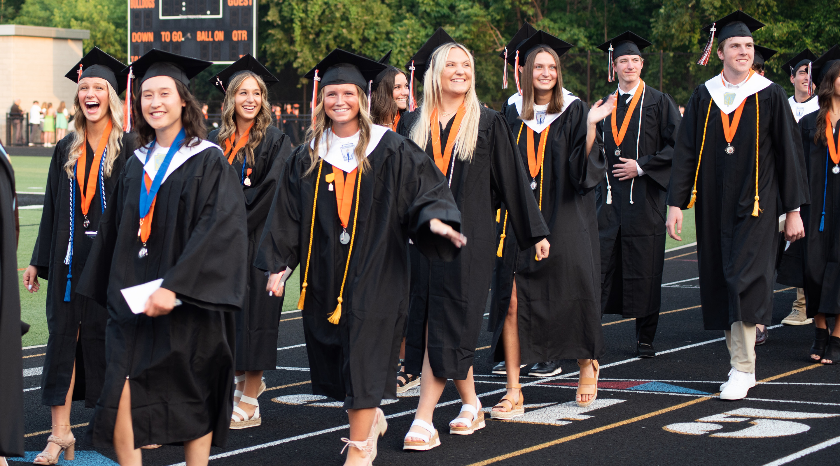 Group of smiling graduates wearing black caps and gowns walking on a track during an outdoor ceremony.