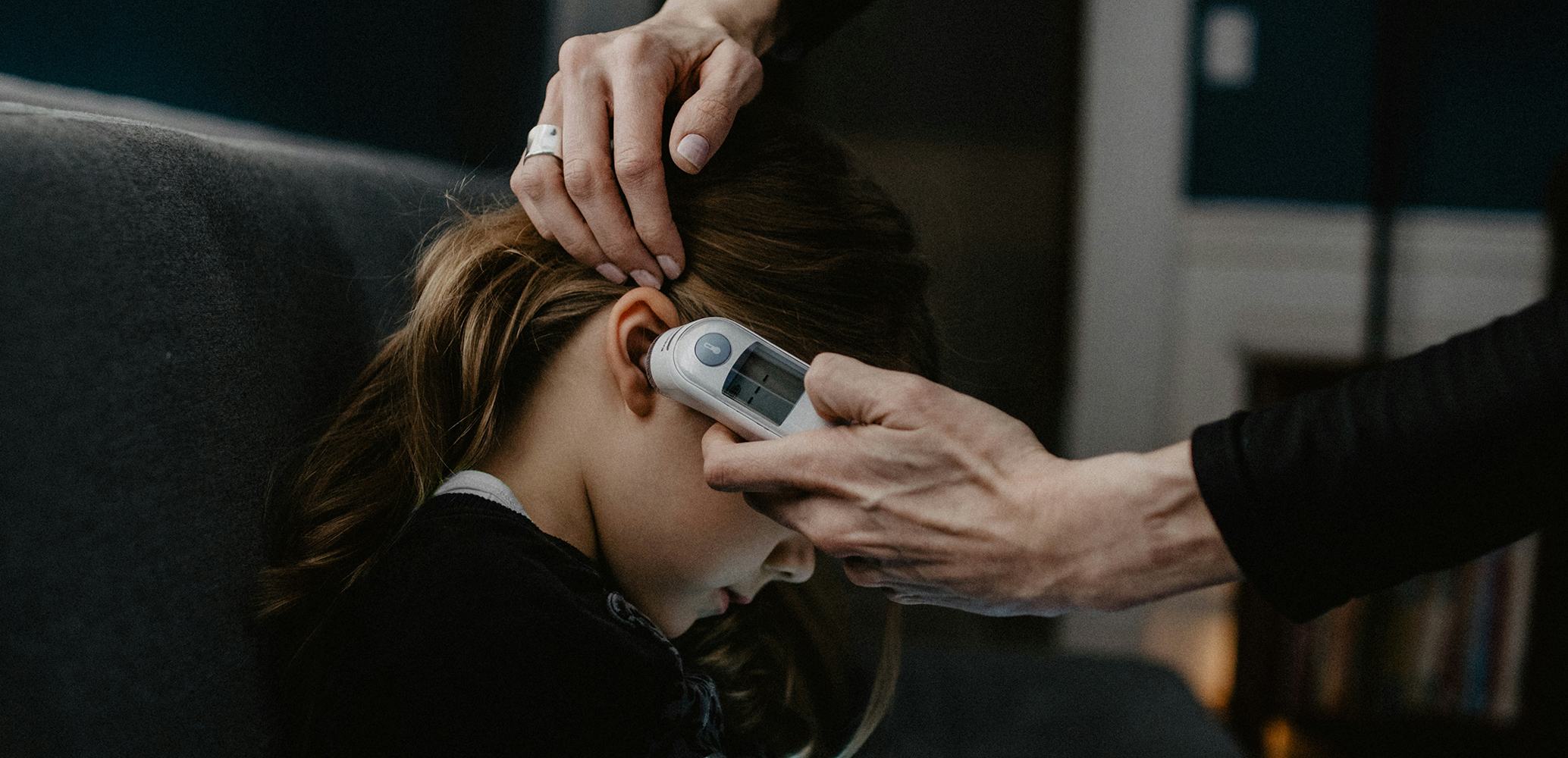 Adult using an ear thermometer to measure the temperature of a child sitting on a couch.